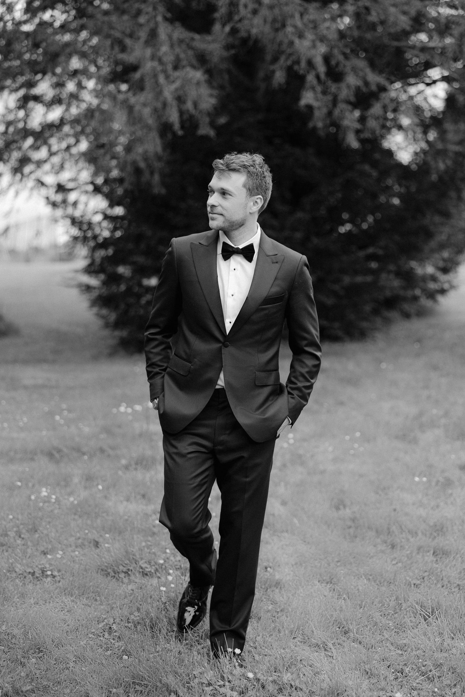 Groom in black tuxedo with bow tie walks confidently outdoors in black and white wedding photo.