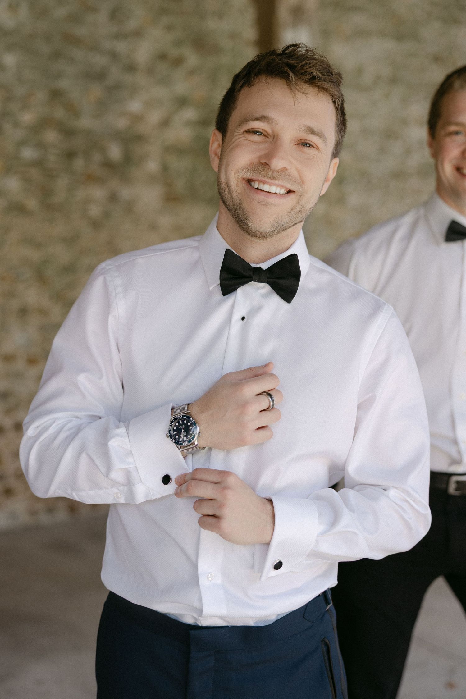 Smiling groom in white tuxedo shirt and black bow tie adjusting cufflinks before wedding ceremony.
