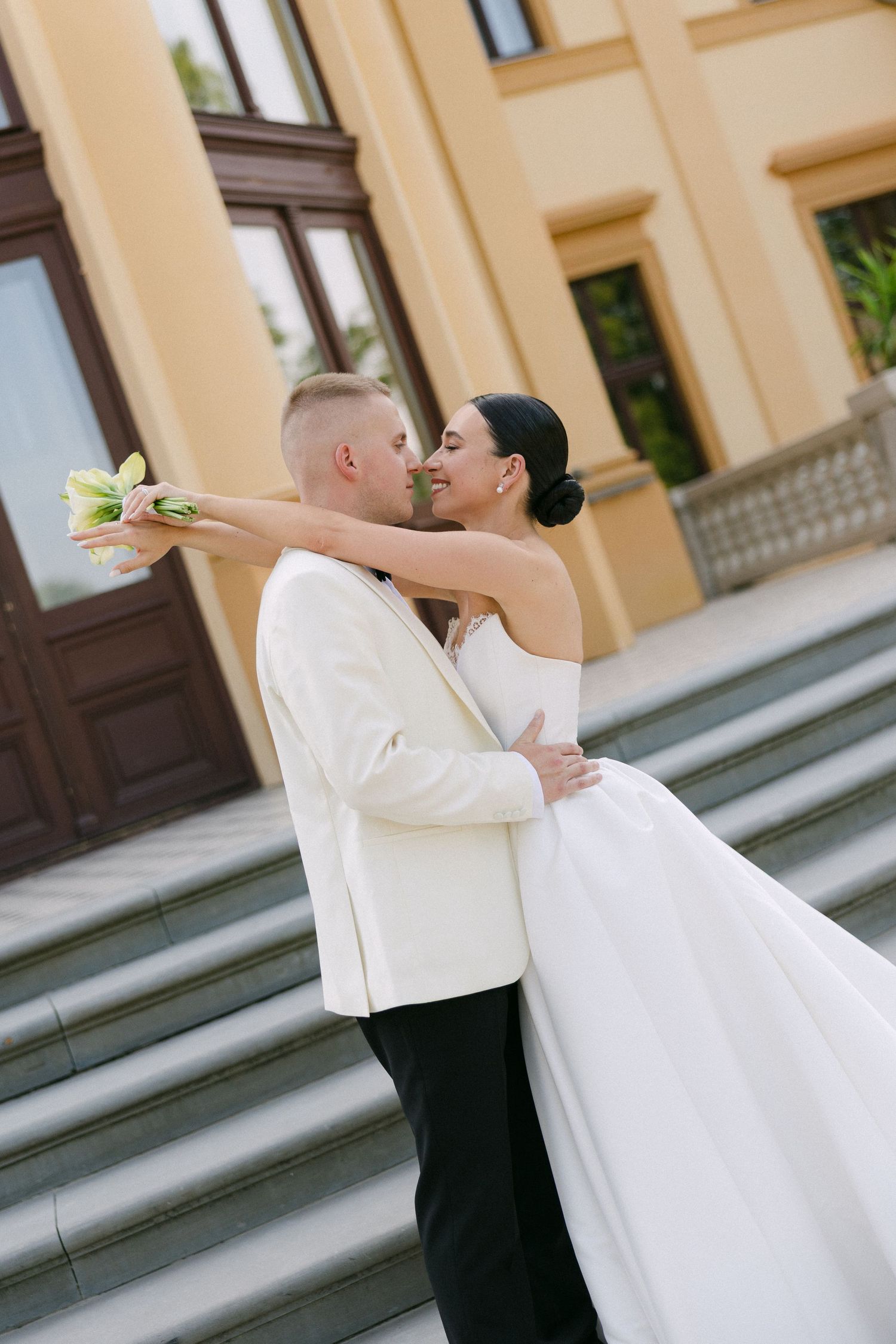 Bride and groom sharing a romantic embrace outside a yellow building, bride holding white tulip bouquet.