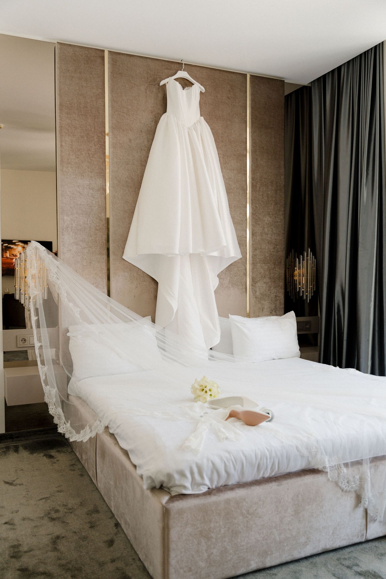 White wedding dress hanging above a bed with veil, bouquet, and bridal shoes in a luxury hotel room.