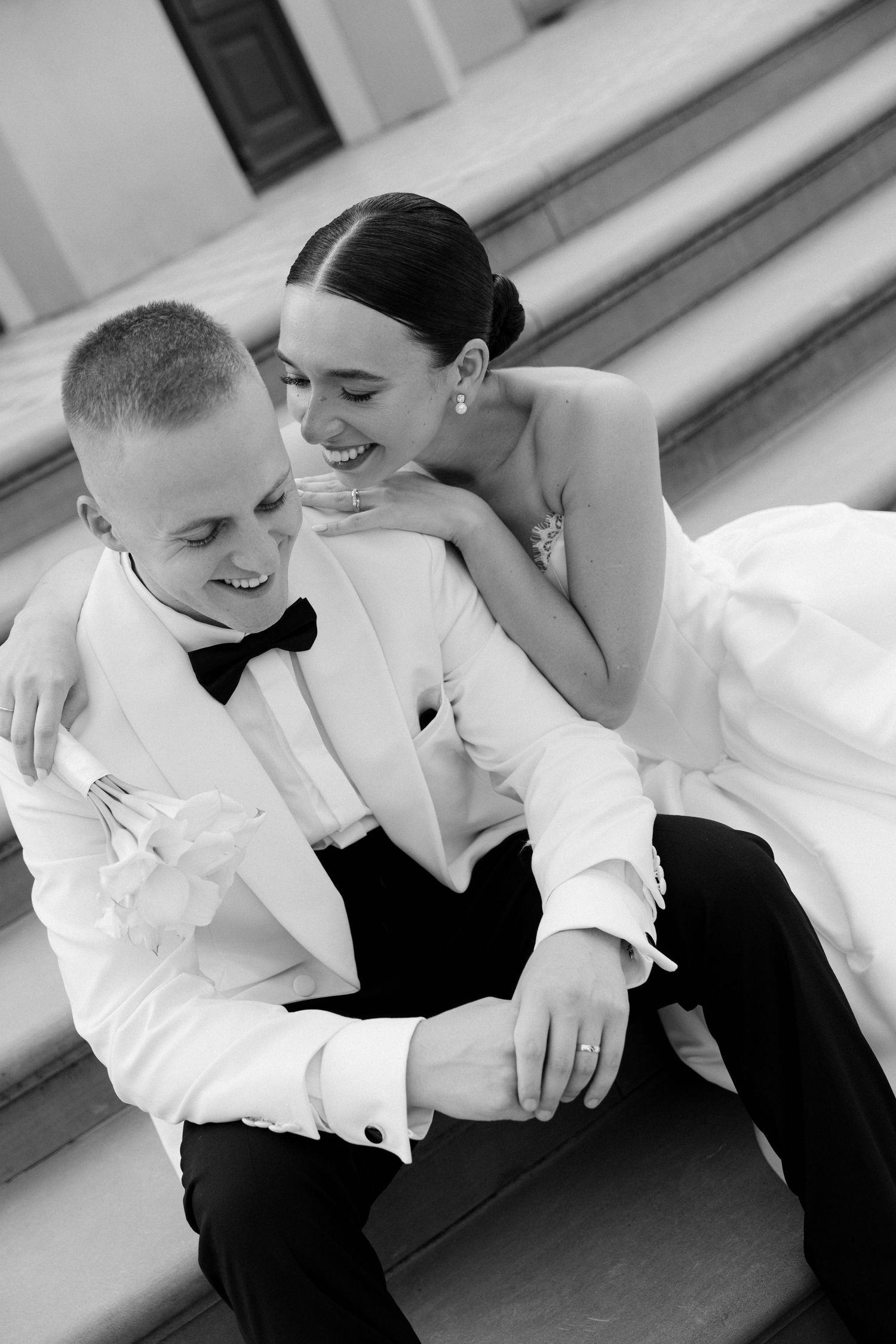 Black and white photo of joyful bride and groom sitting on steps laughing together on their wedding day.