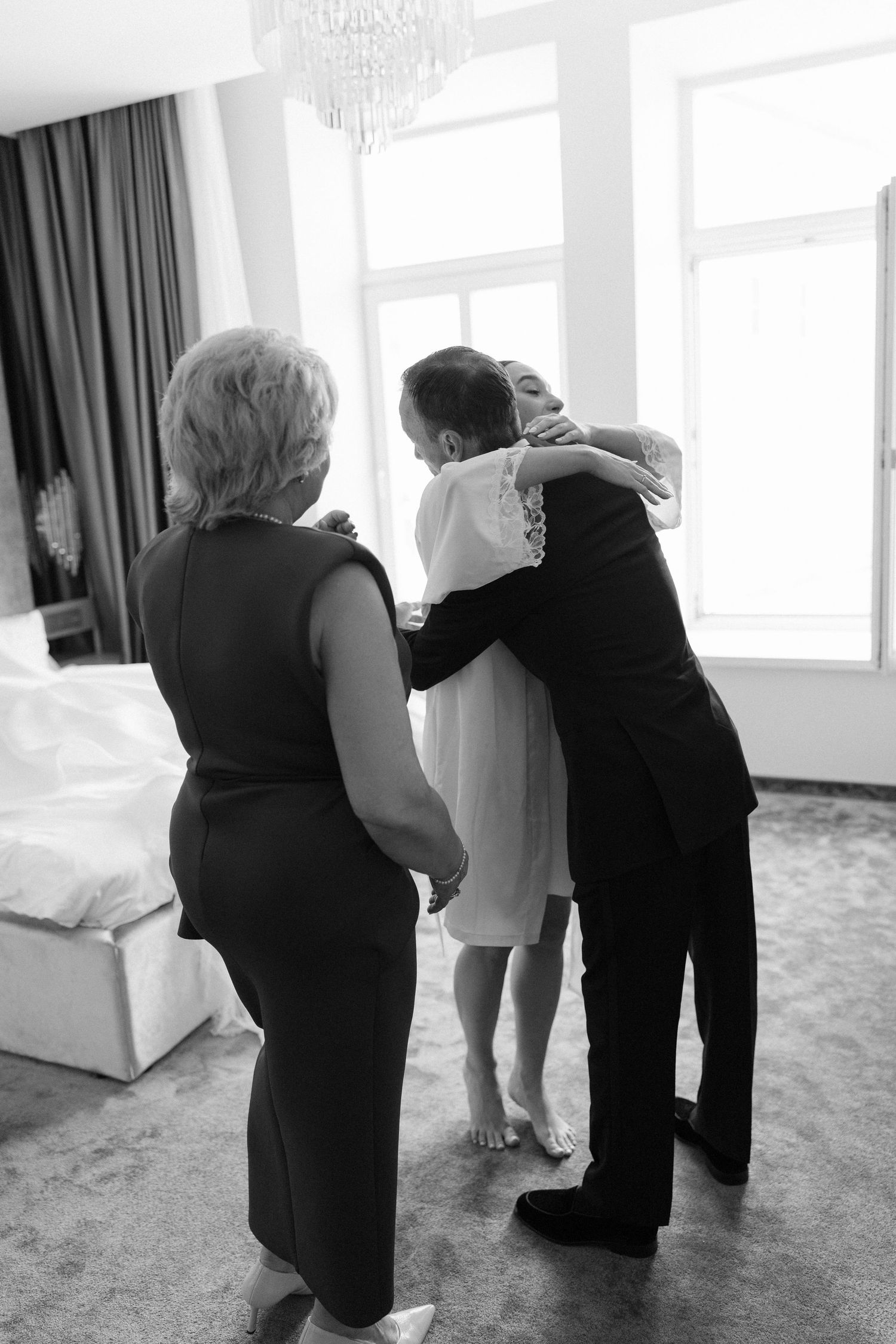 Black and white photo of a groom hugging bride while mother watches in elegant hotel room.