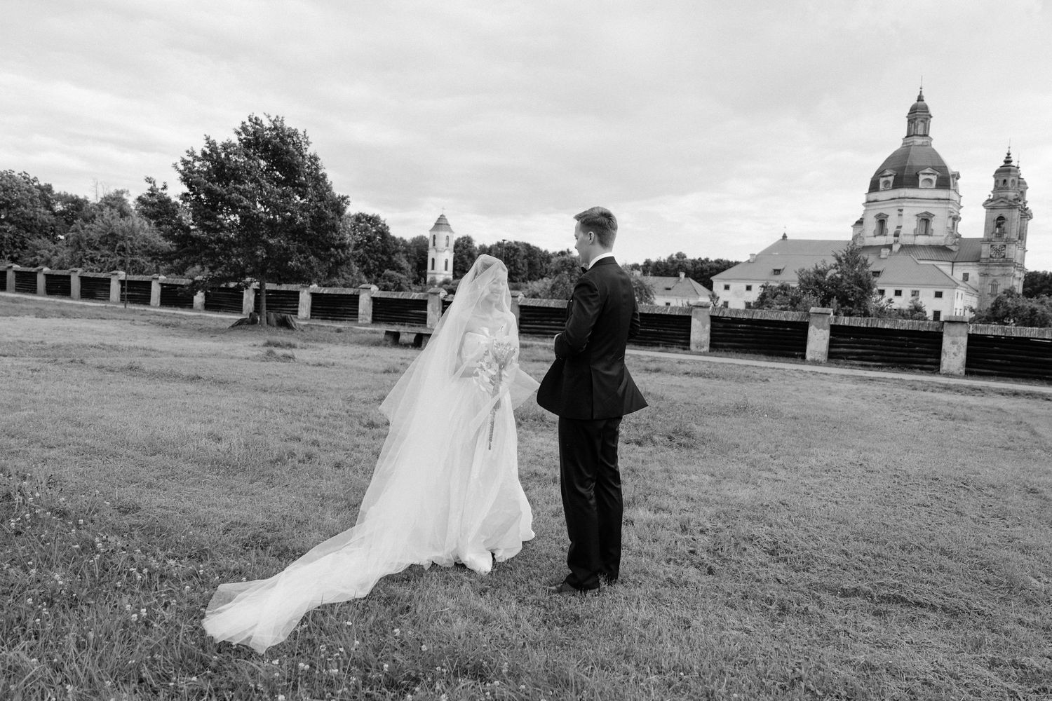 Black and white photo of a bride and groom walking in a field with a historic church in the background.