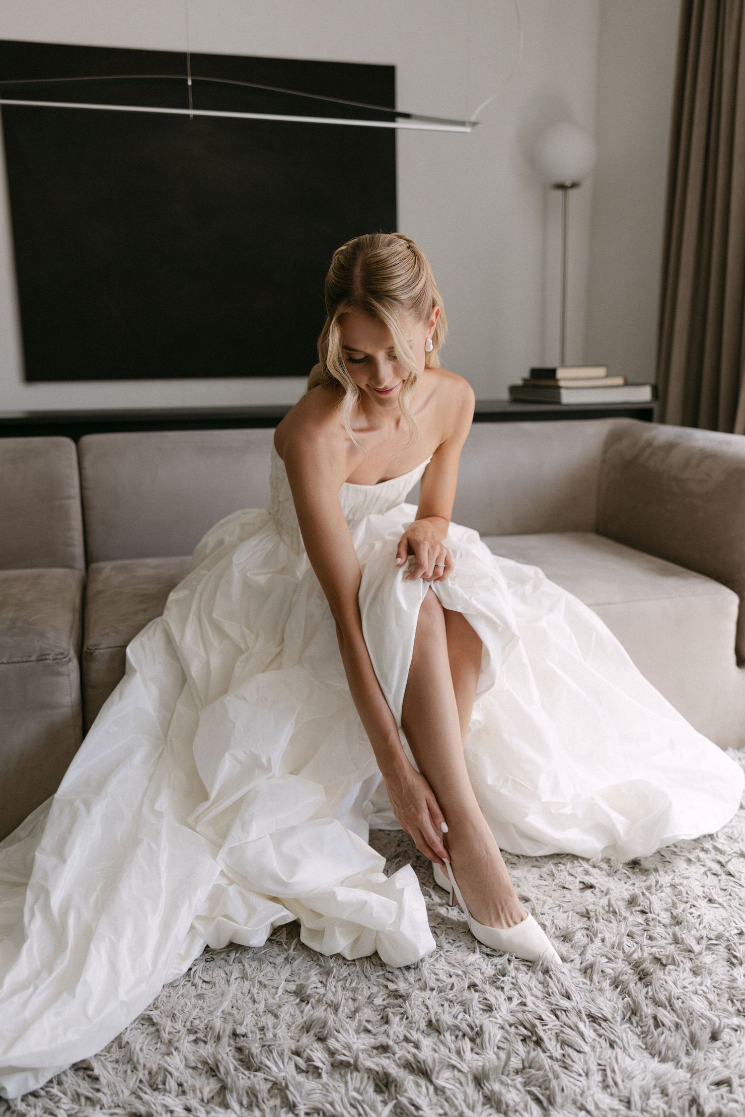 Bride in a white ball gown sits on a sofa adjusting her white heels on a plush carpet.