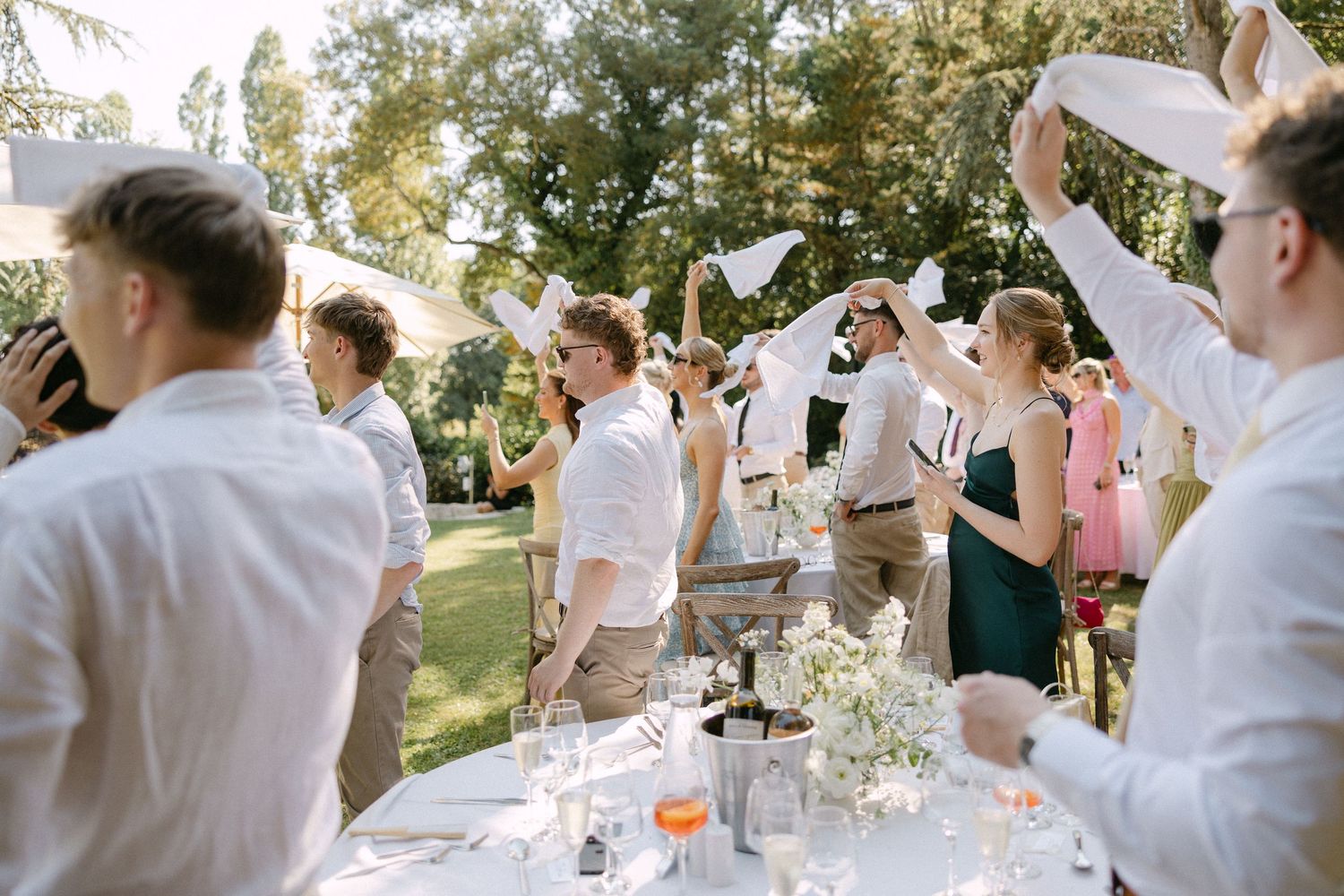Wedding guests waving white napkins in celebration at an outdoor garden reception on a sunny day.