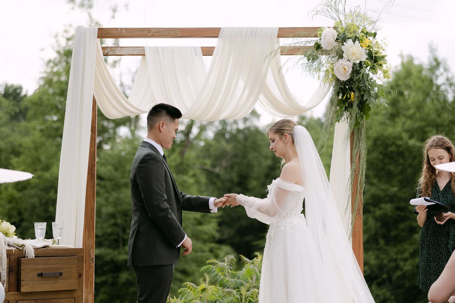 A bride and groom exchange vows under a floral wooden arch at an outdoor wedding ceremony.