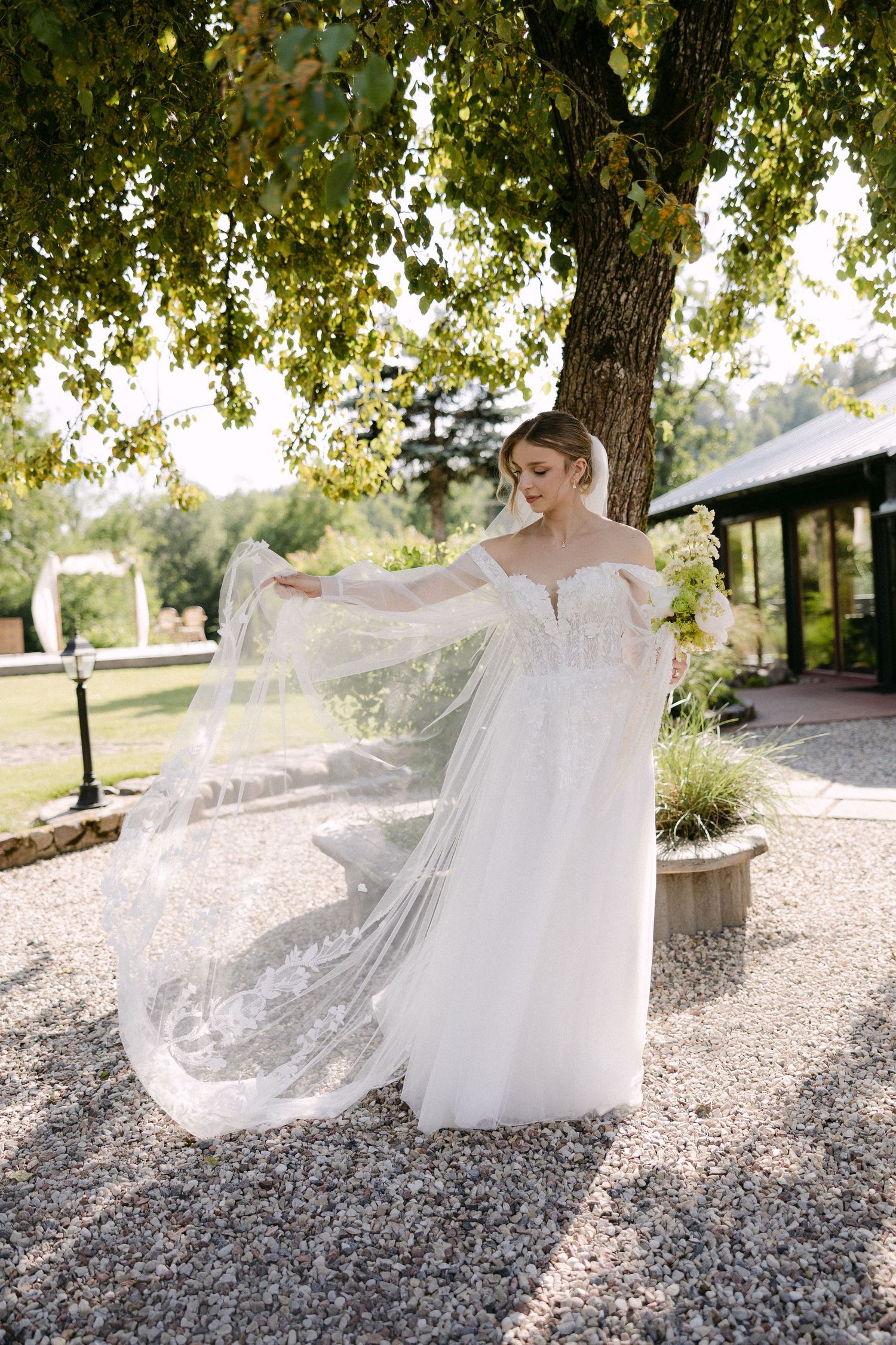 Bride in lace off-shoulder gown twirls flowing veil under a large tree in garden setting.