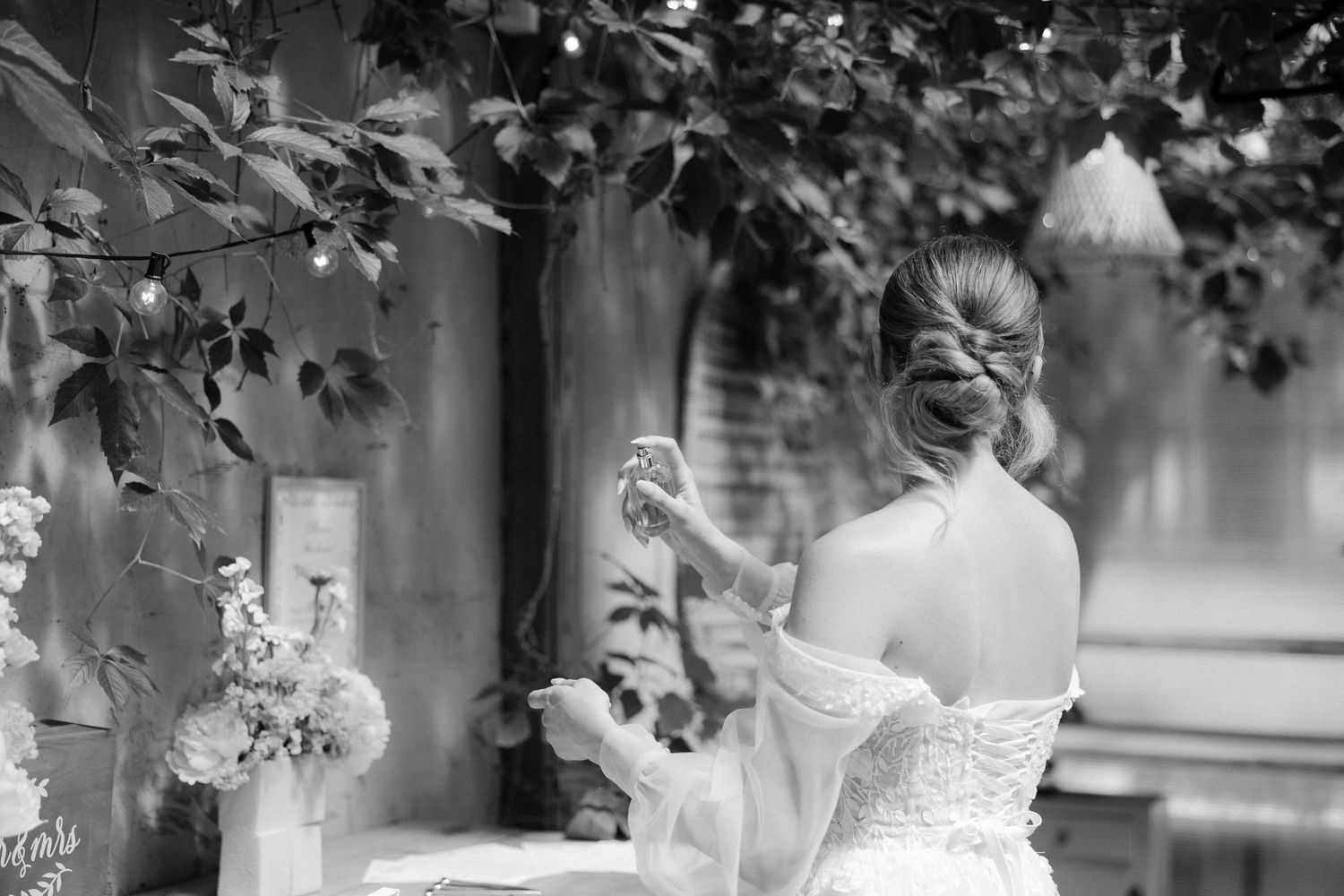 Bride in elegant off-shoulder gown with updo hairstyle seated outdoors among lush greenery in black and white.