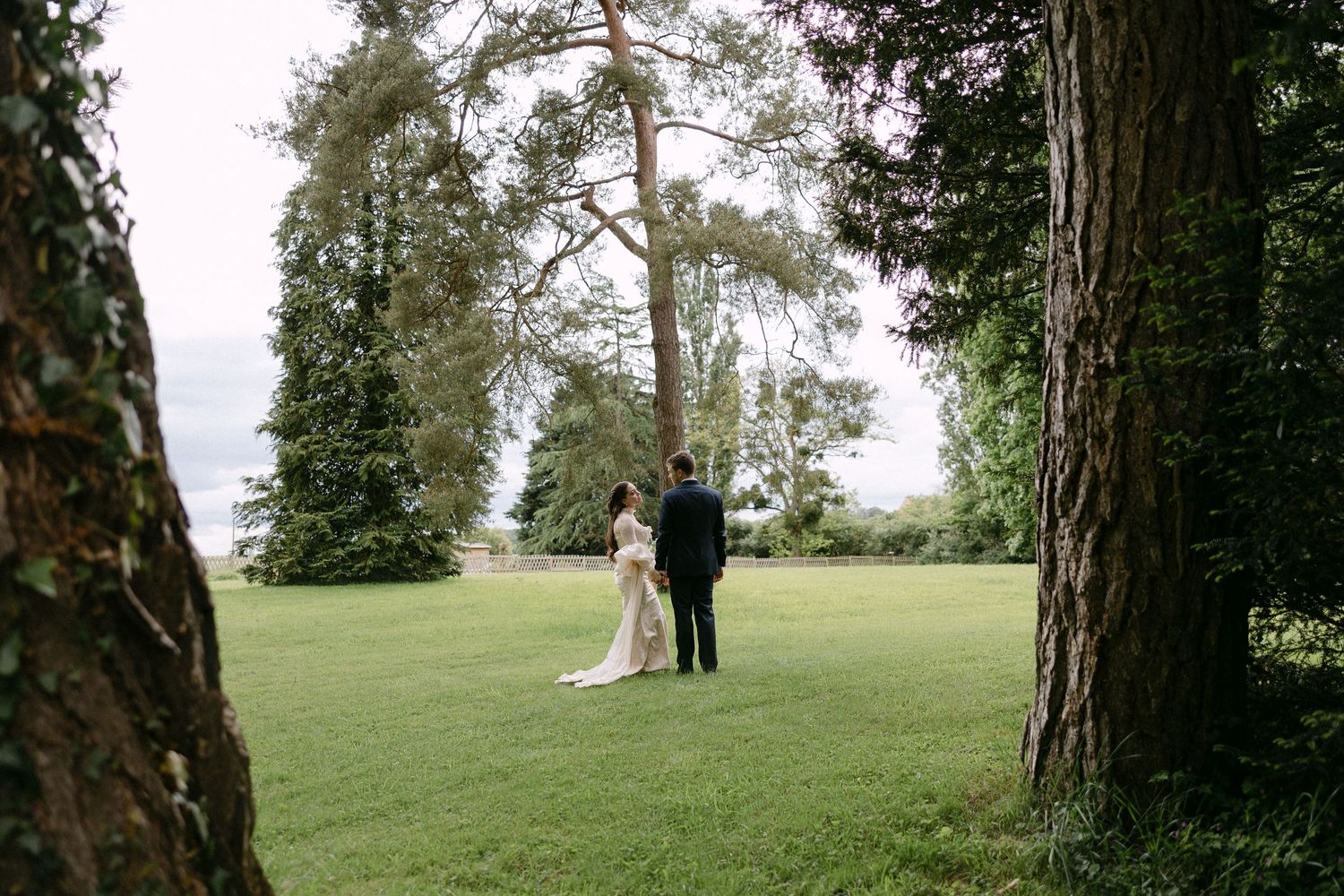 Bride and groom walking hand in hand across lush green lawn framed by tall trees at outdoor wedding.