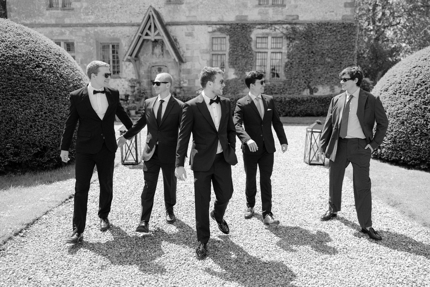 Five groomsmen in black tuxedos walking confidently together outside a grand estate in a black and white wedding photo.