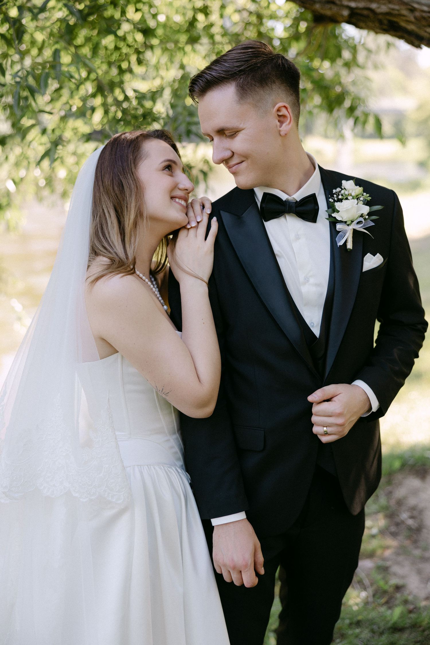 Smiling bride in white dress leaning on groom in black tuxedo with bow tie outdoors.