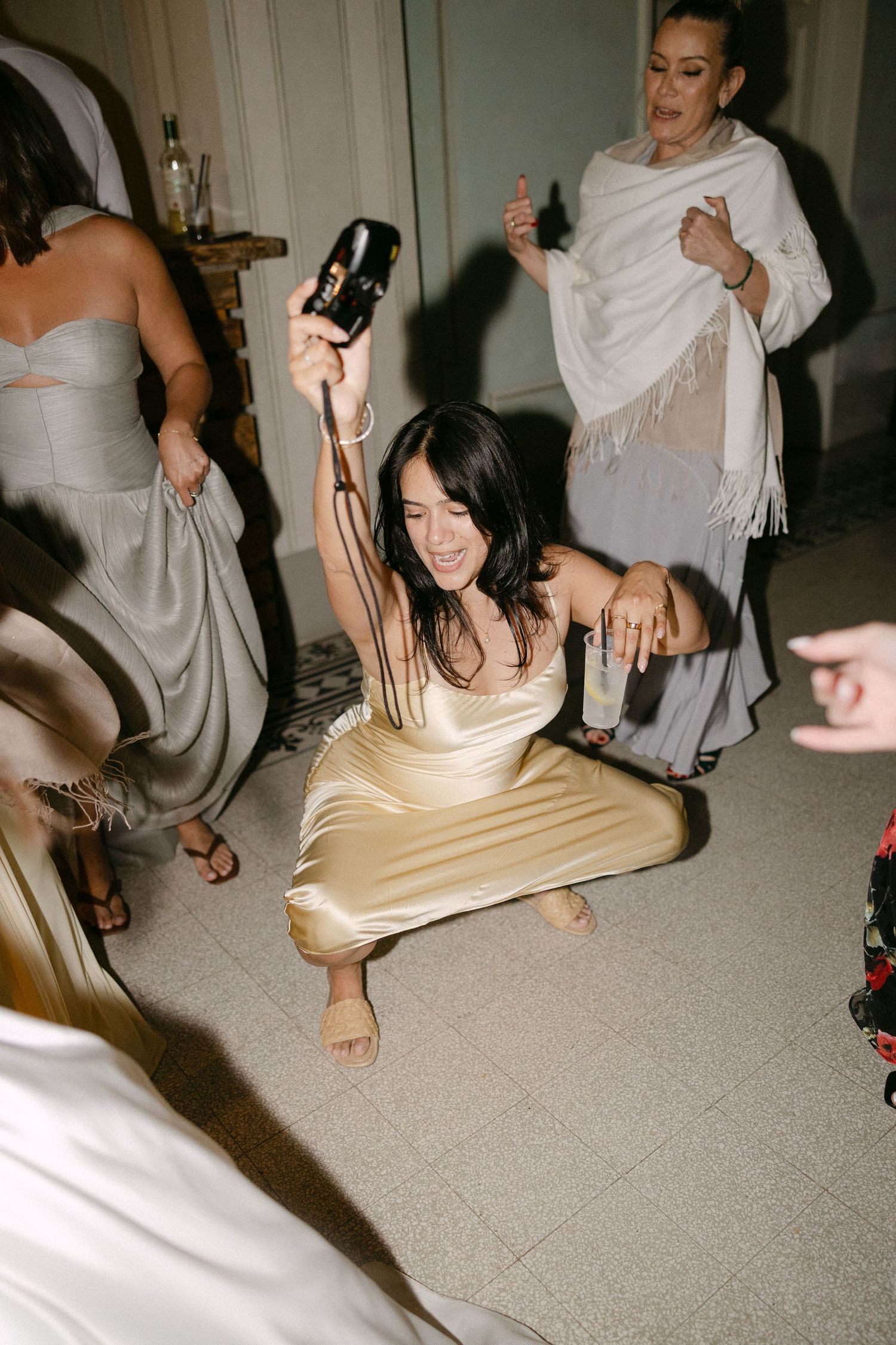 Bridesmaid in gold satin dress crouches low on dance floor holding camera aloft at wedding reception.