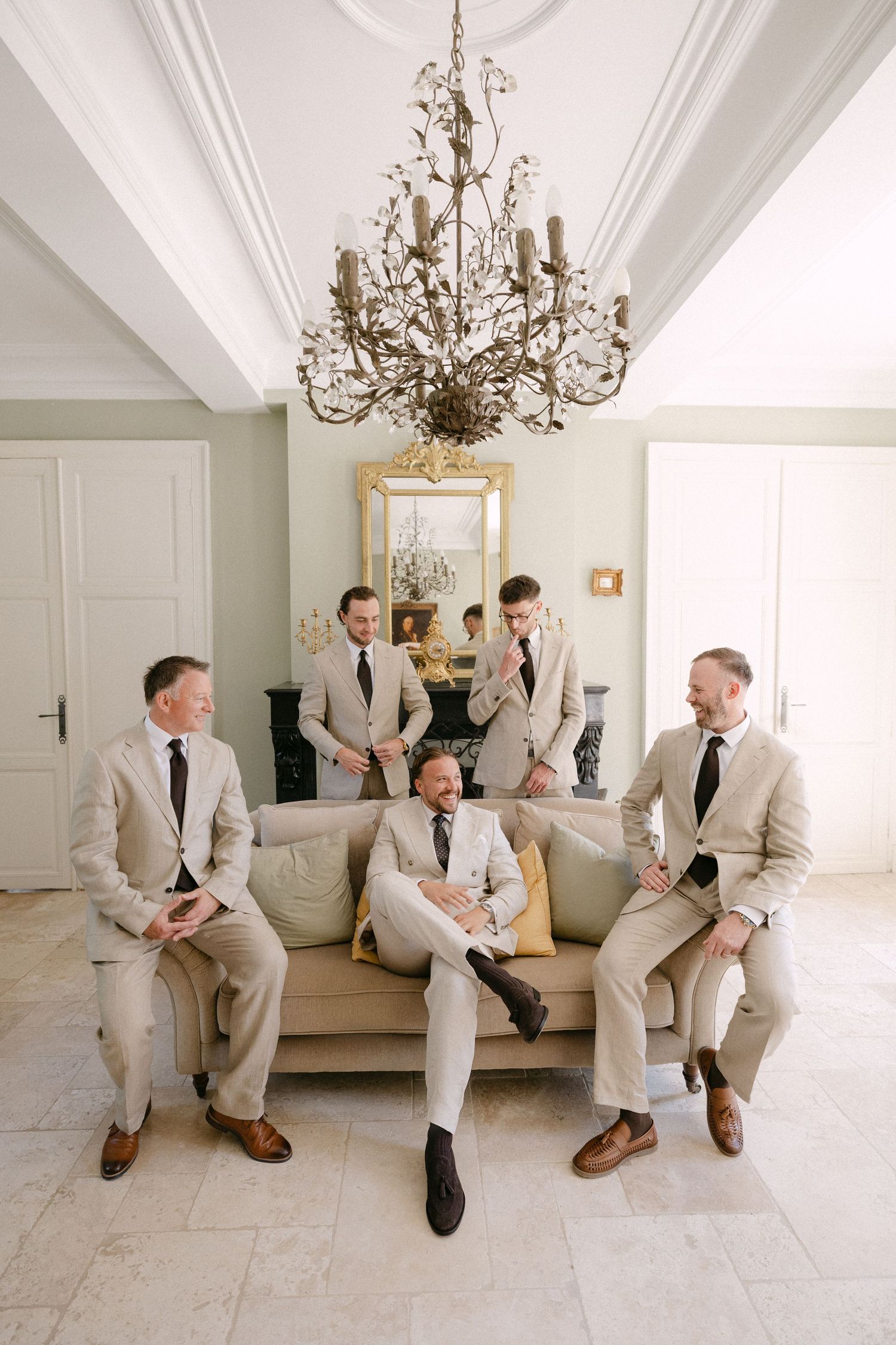Five groomsmen in matching tan suits and black ties pose elegantly in a bright room with a crystal chandelier.