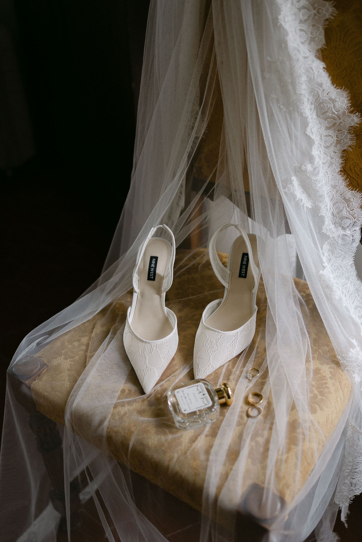 White bridal heels, perfume bottle, and gold rings displayed on a chair draped with an elegant lace wedding veil.