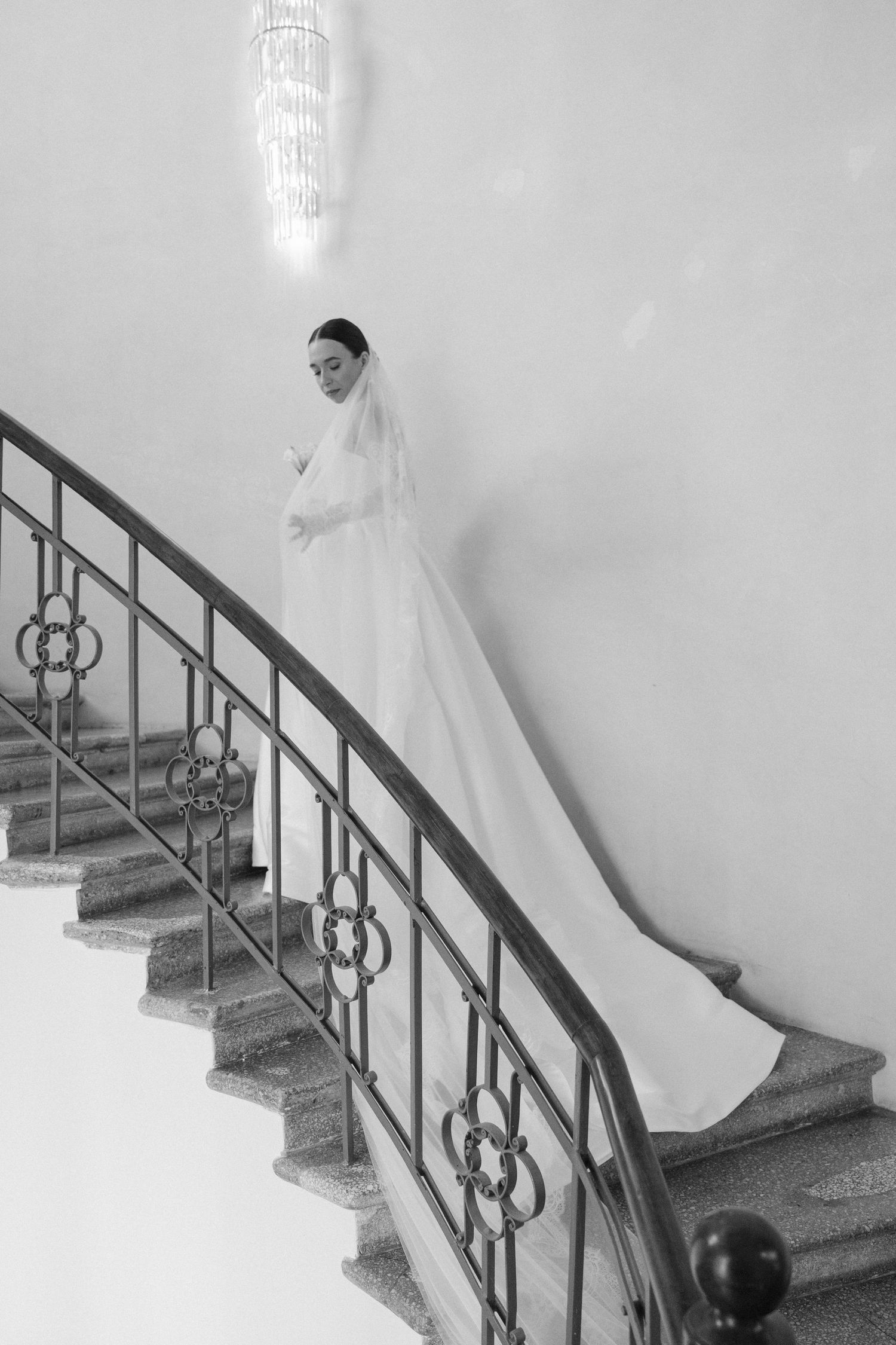 Elegant bride in white gown and long veil descending a curved staircase in black and white photo.