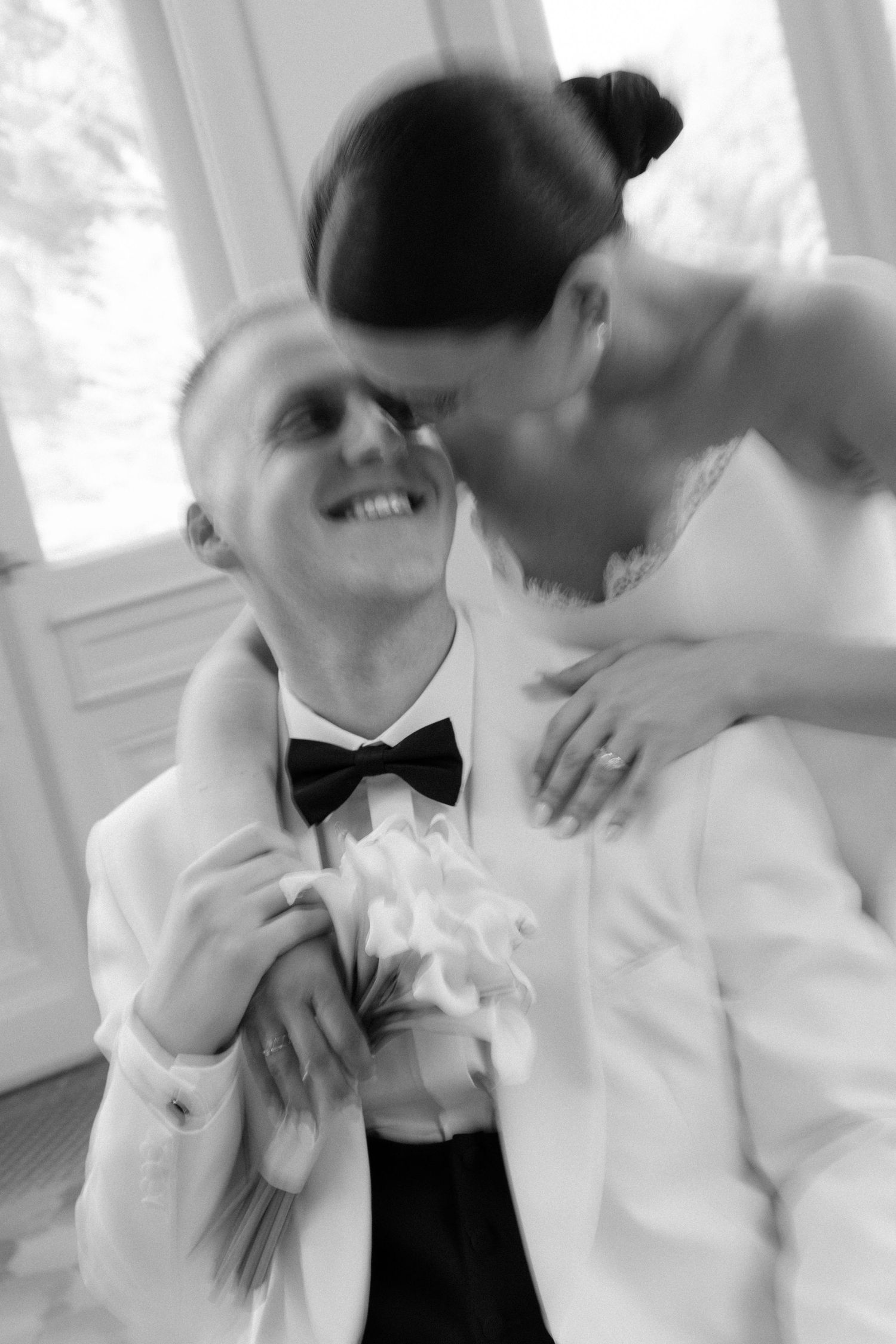 Black and white photo of smiling groom in white tuxedo embraced by bride holding calla lily bouquet.
