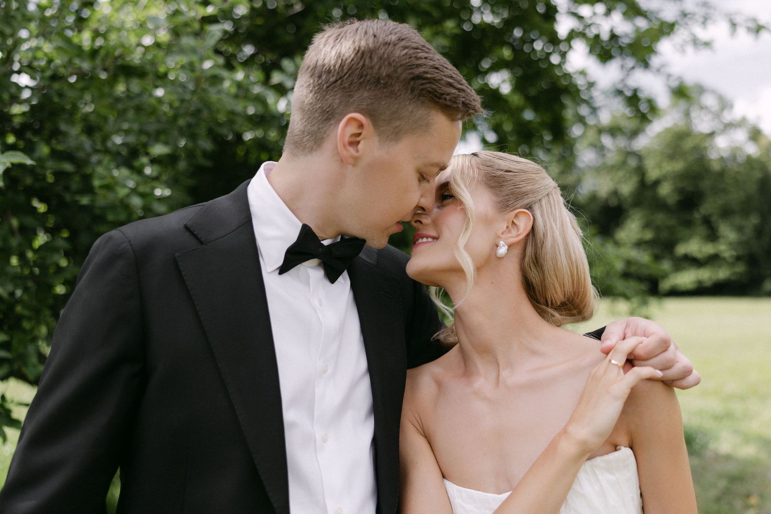 Bride and groom sharing a romantic nose-to-nose moment outdoors surrounded by lush green trees.