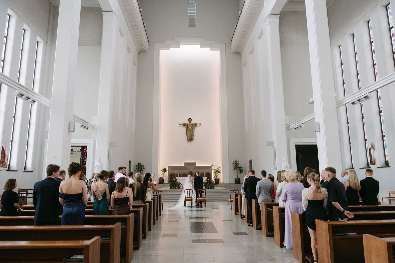 Guests seated in wooden pews during a wedding ceremony inside a modern white church with a crucifix above the altar.