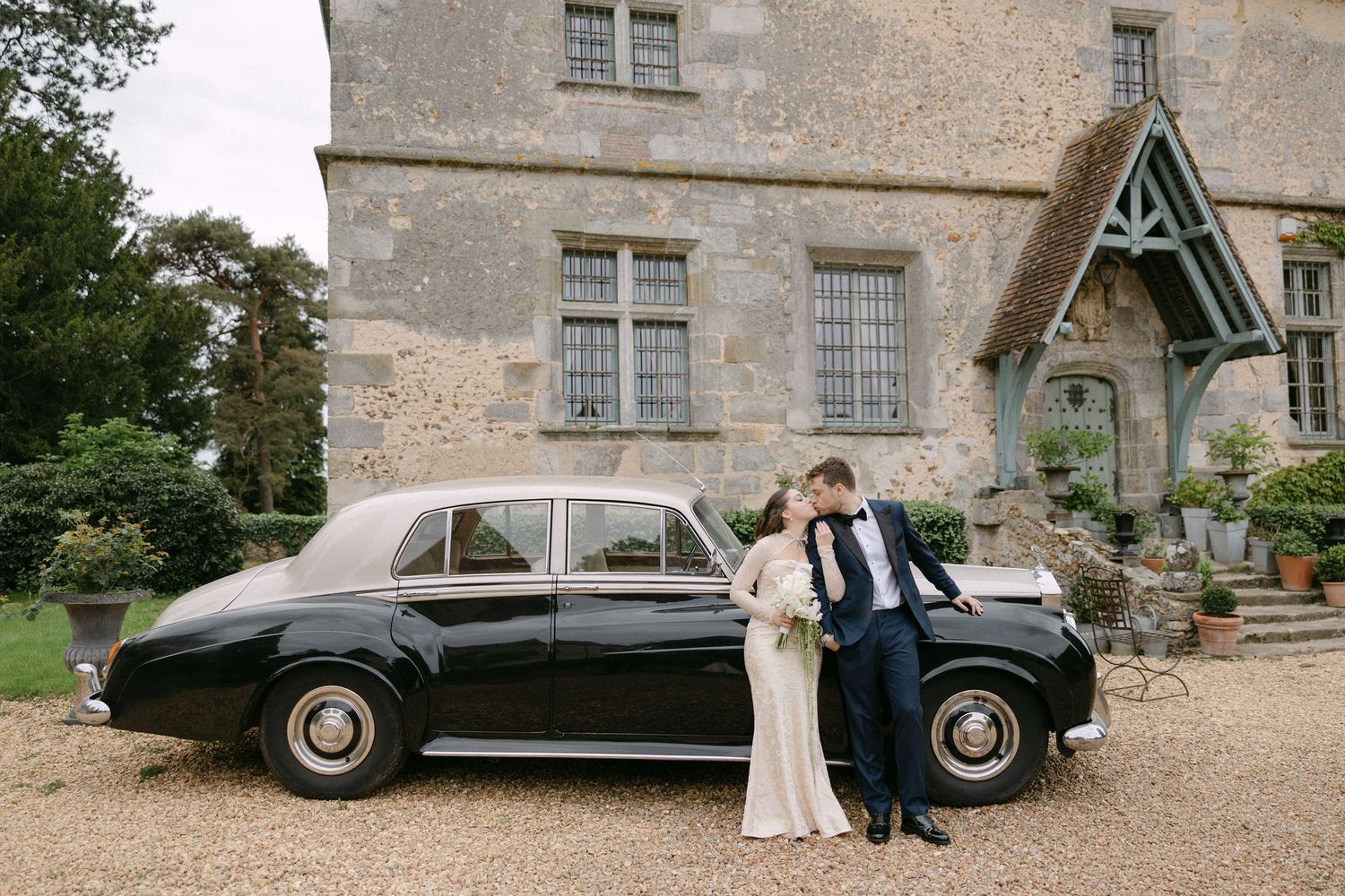 Bride and groom pose beside a classic black Rolls-Royce outside a historic stone manor house.