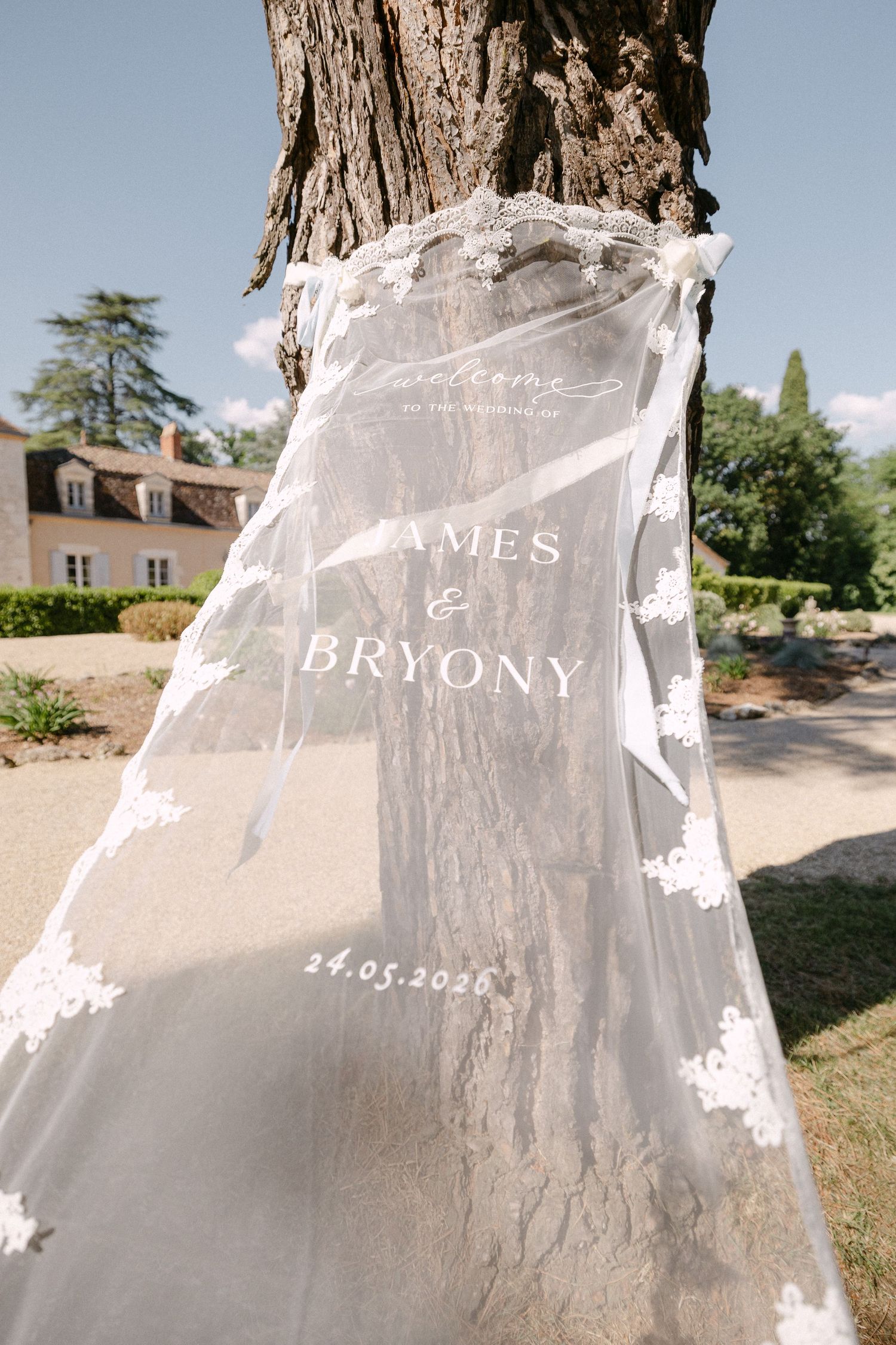A sheer lace wedding veil tied to a tree displaying James and Bryony wedding welcome sign dated 24.05.2025.