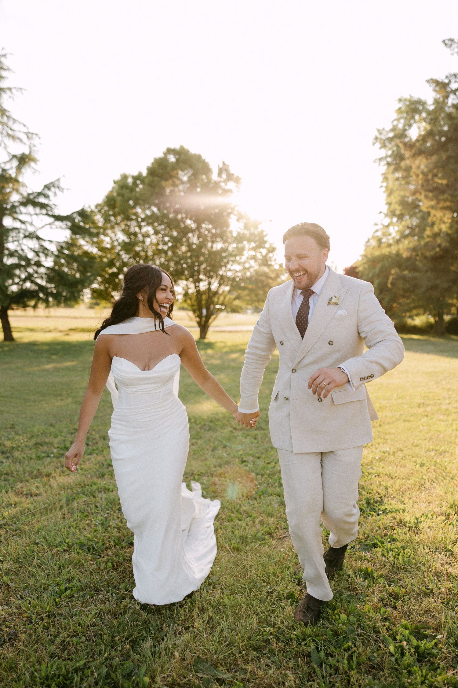 Bride and groom walk hand-in-hand laughing across sunlit green field at golden hour.