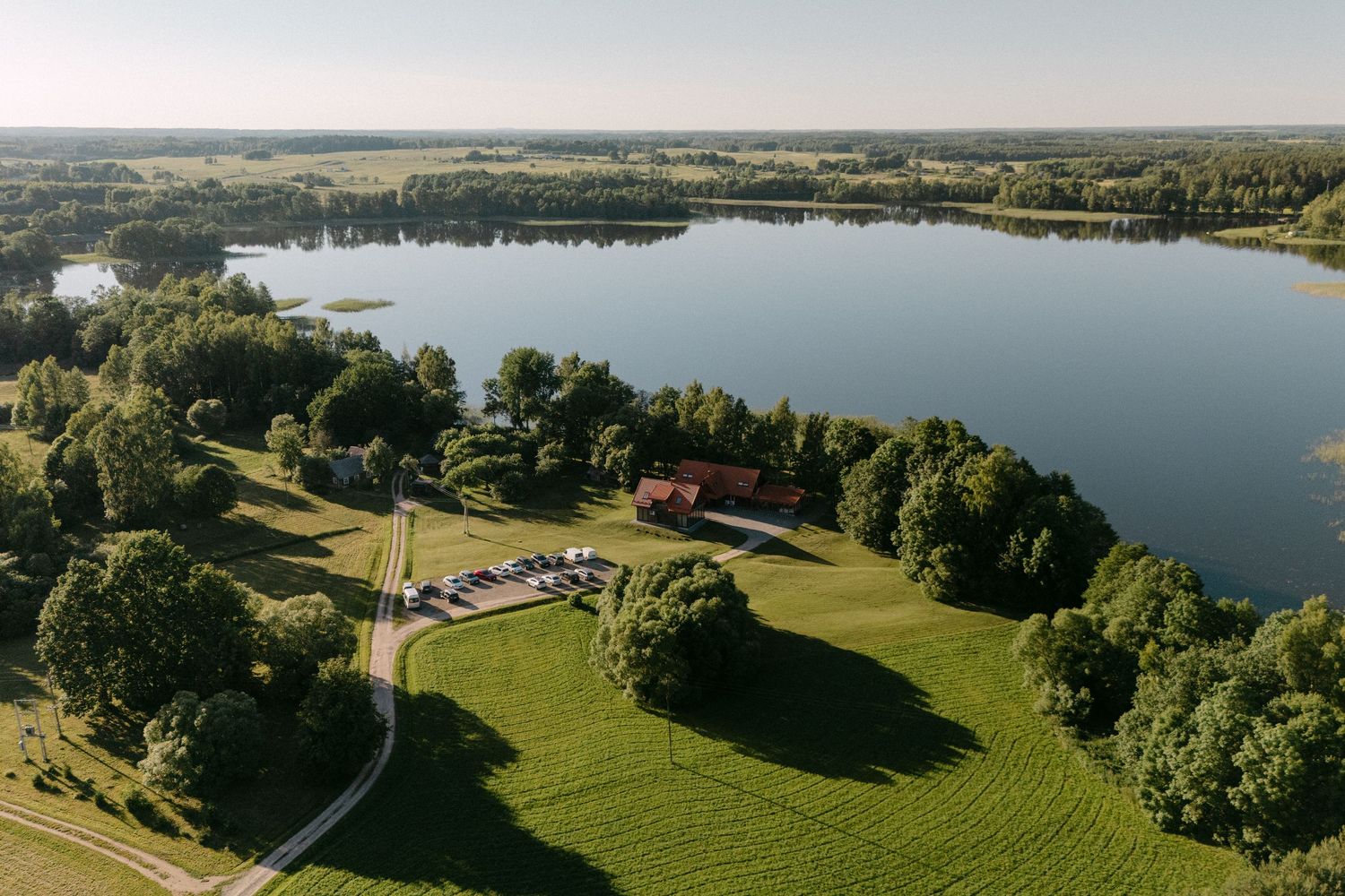 Aerial view of a scenic lakeside venue surrounded by lush green trees and open fields on a sunny day.