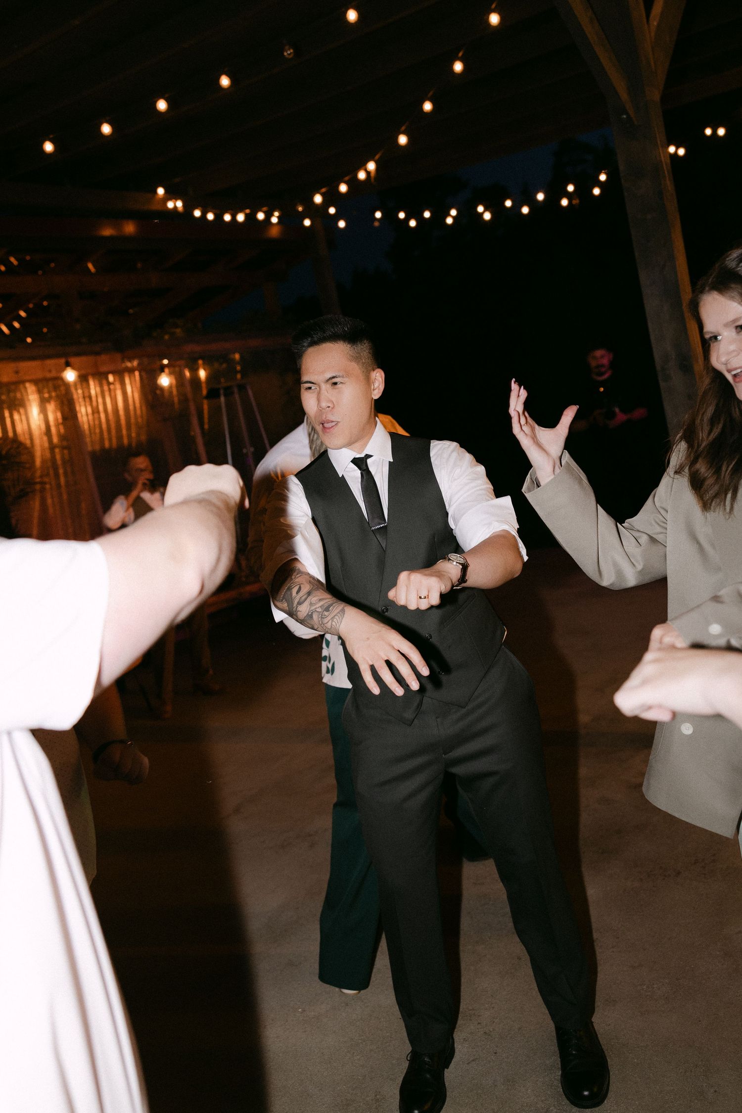 Groom in vest and tie dancing energetically with guests at outdoor wedding reception at night.