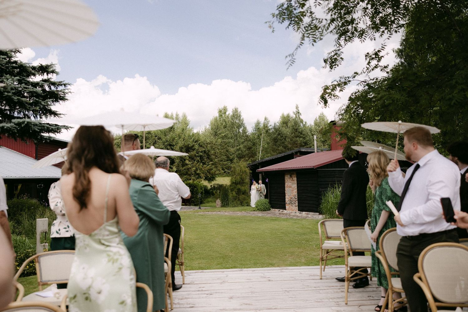 Outdoor wedding ceremony on a wooden deck with guests seated in chairs, lush green lawn and trees in the background.