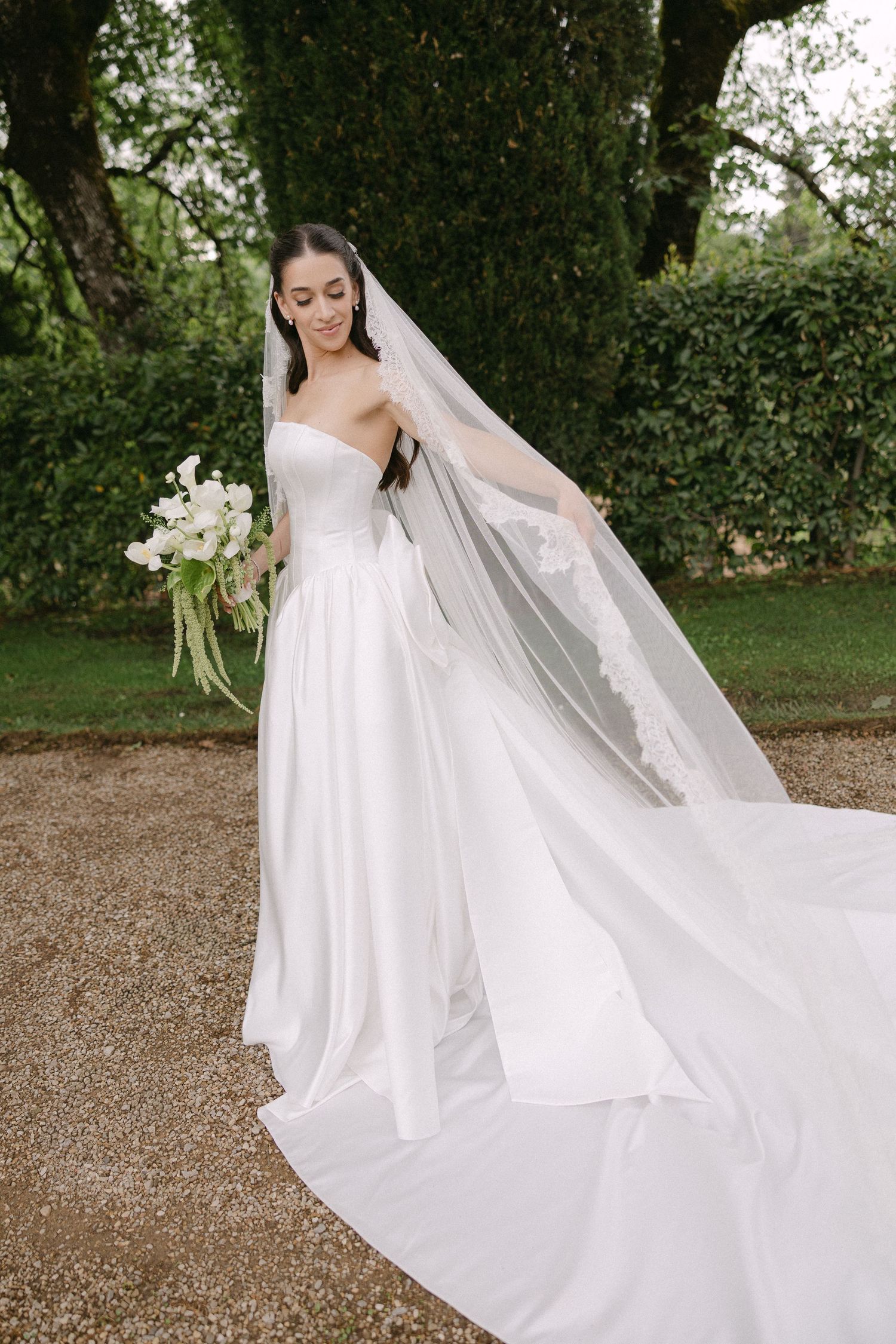 Bride in strapless white ball gown with cathedral veil holds bouquet in lush garden setting.