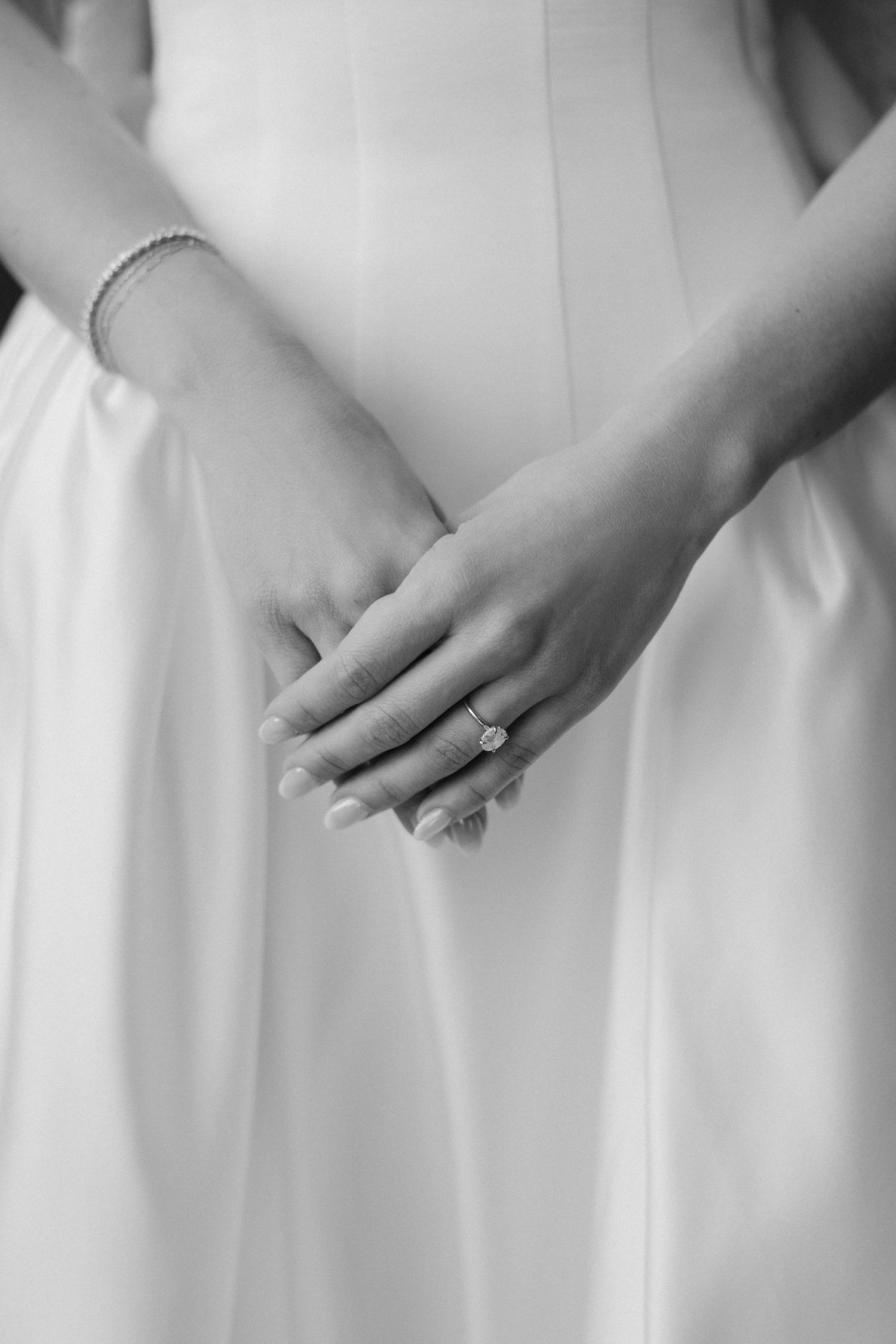 Black and white close-up of bride's hands showing engagement ring and pearl bracelet against white wedding gown.