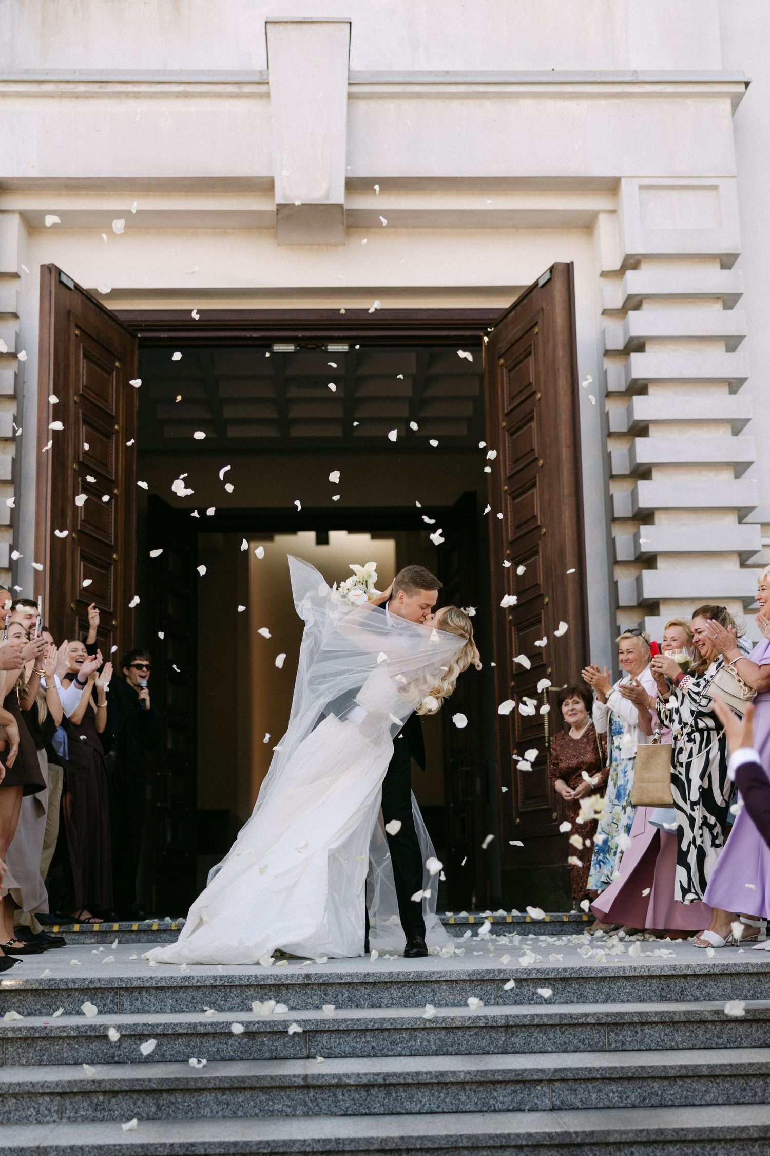 Bride and groom share a romantic dip kiss on church steps while guests celebrate with white confetti.