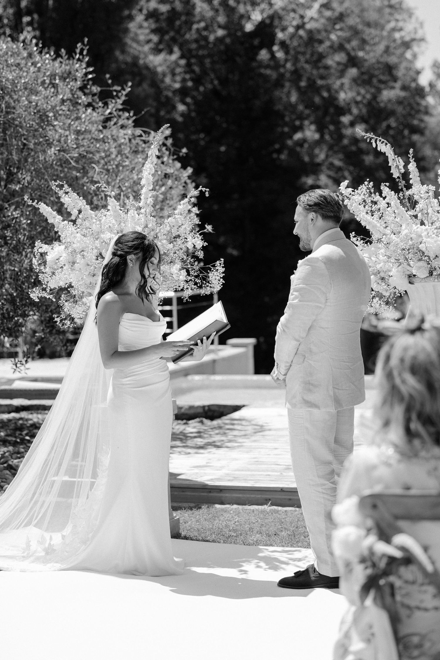 Black and white photo of bride reading vows to groom at outdoor ceremony with floral arch backdrop.