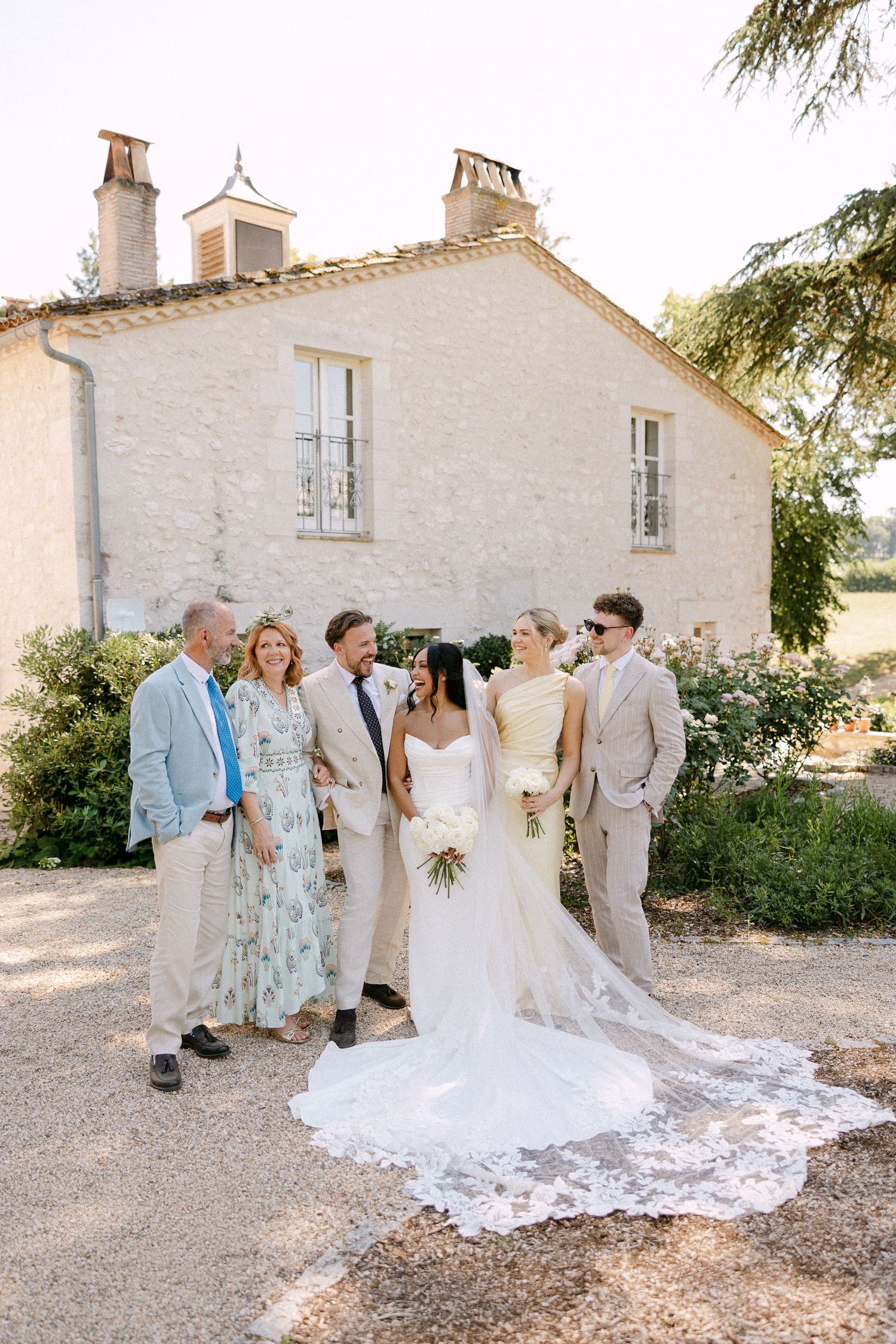 Wedding party poses with bride and groom outside a rustic French stone building on sunny day.