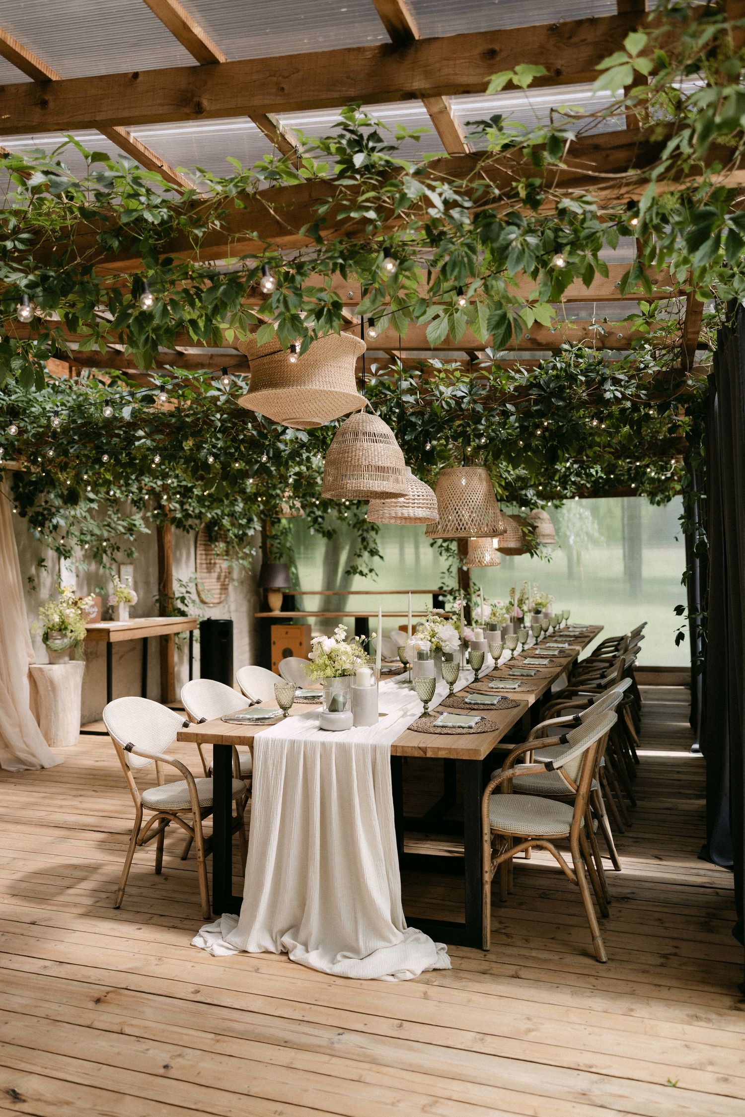 Elegant outdoor wedding reception under a pergola with lush greenery, wicker pendant lights, and a long decorated dining table.
