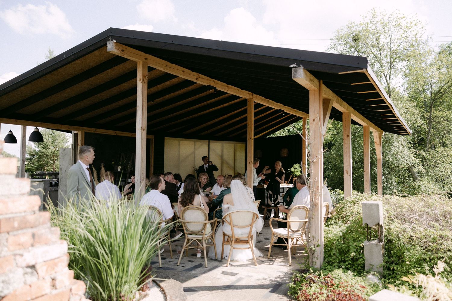 Wedding guests dine under a rustic open-air pavilion surrounded by natural garden greenery.
