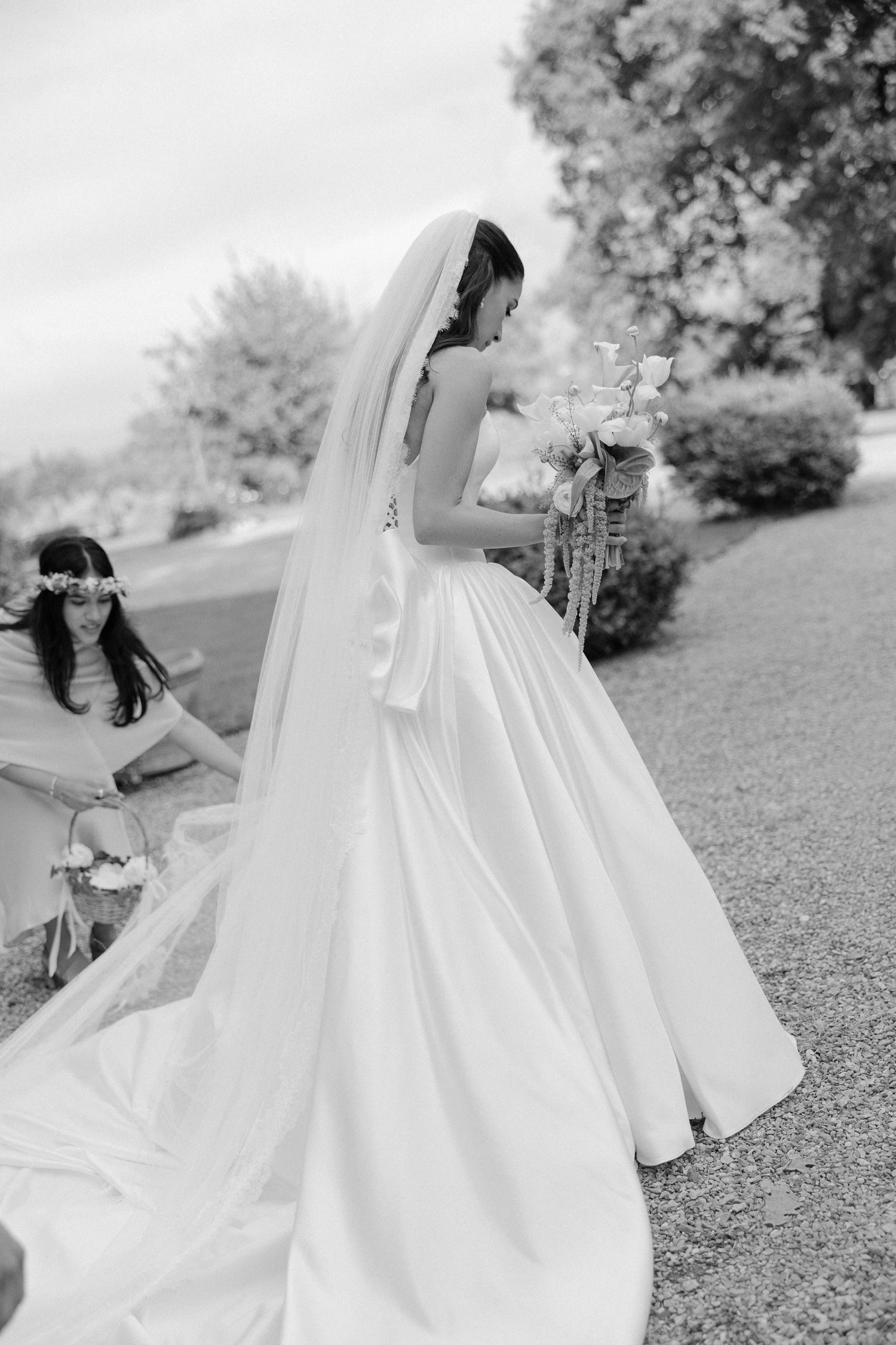 Black and white photo of bride in satin gown holding bouquet while bridesmaid adjusts her train.