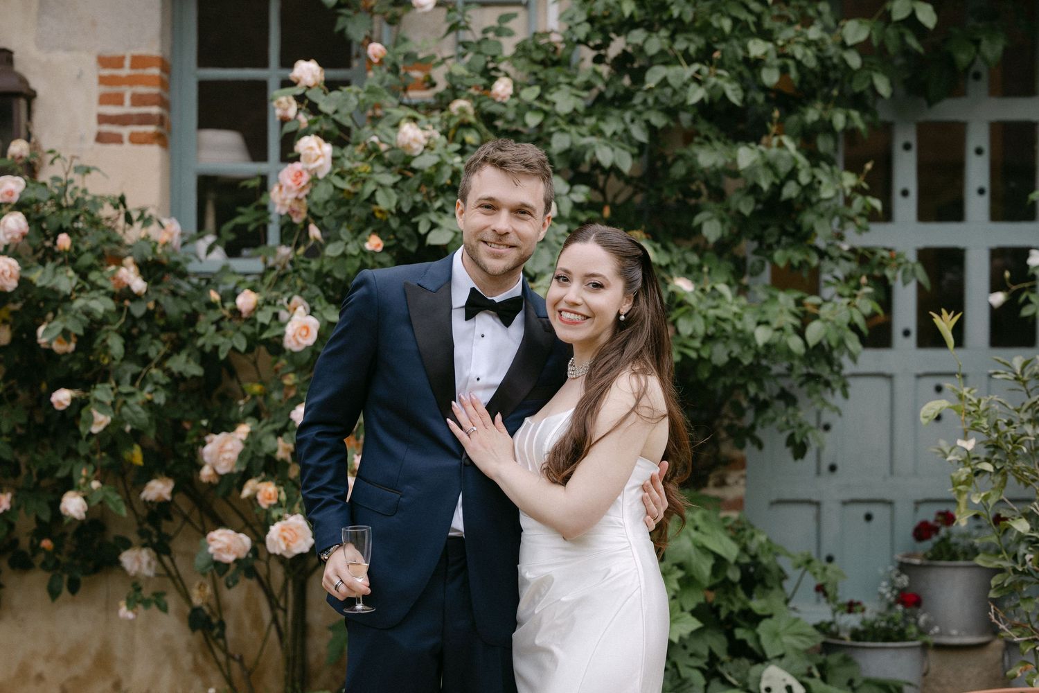 Smiling bride and groom posing together in front of blooming pink rose bushes at outdoor wedding venue.