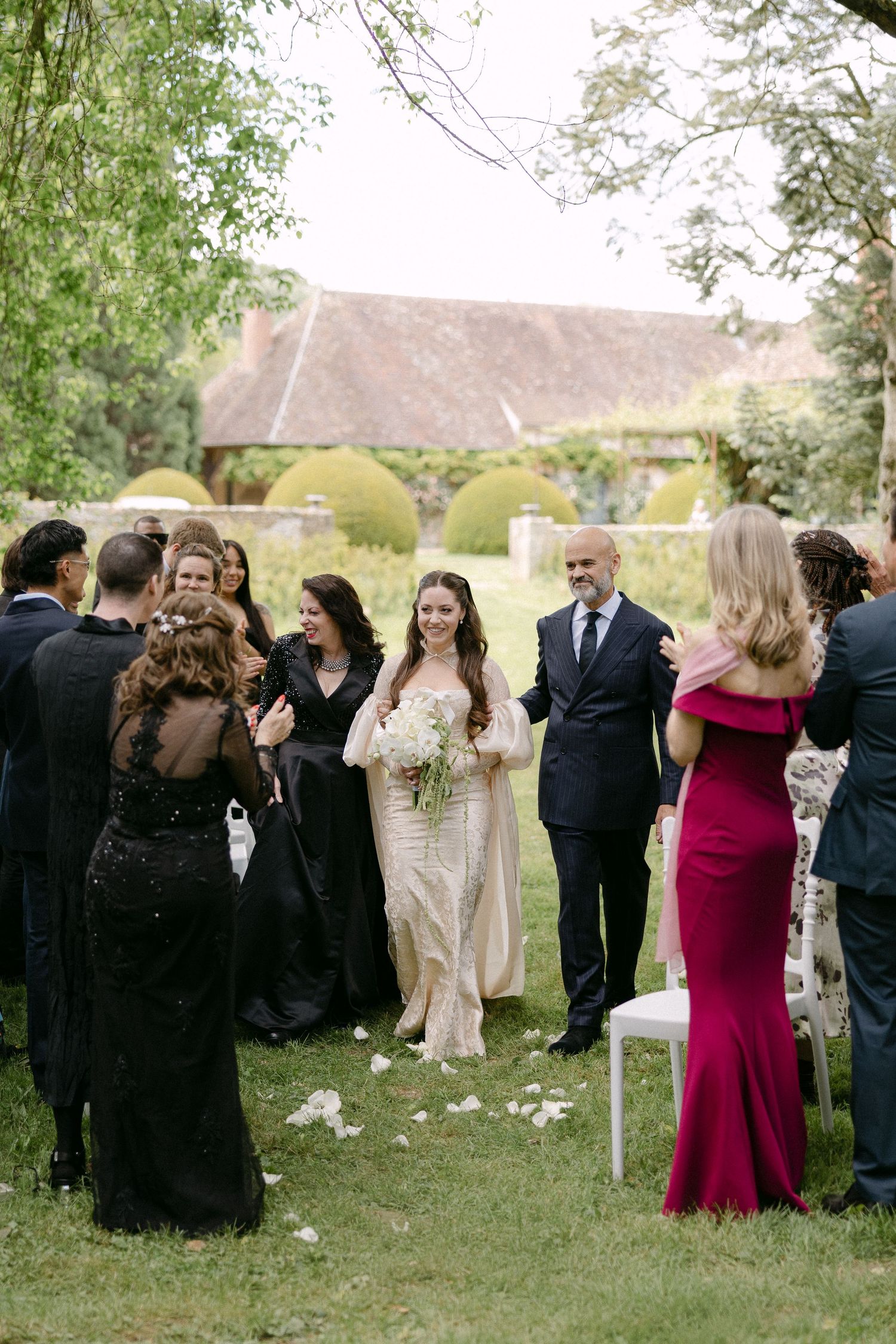 Bride walking down outdoor aisle with father past guests in black formal attire at garden wedding.
