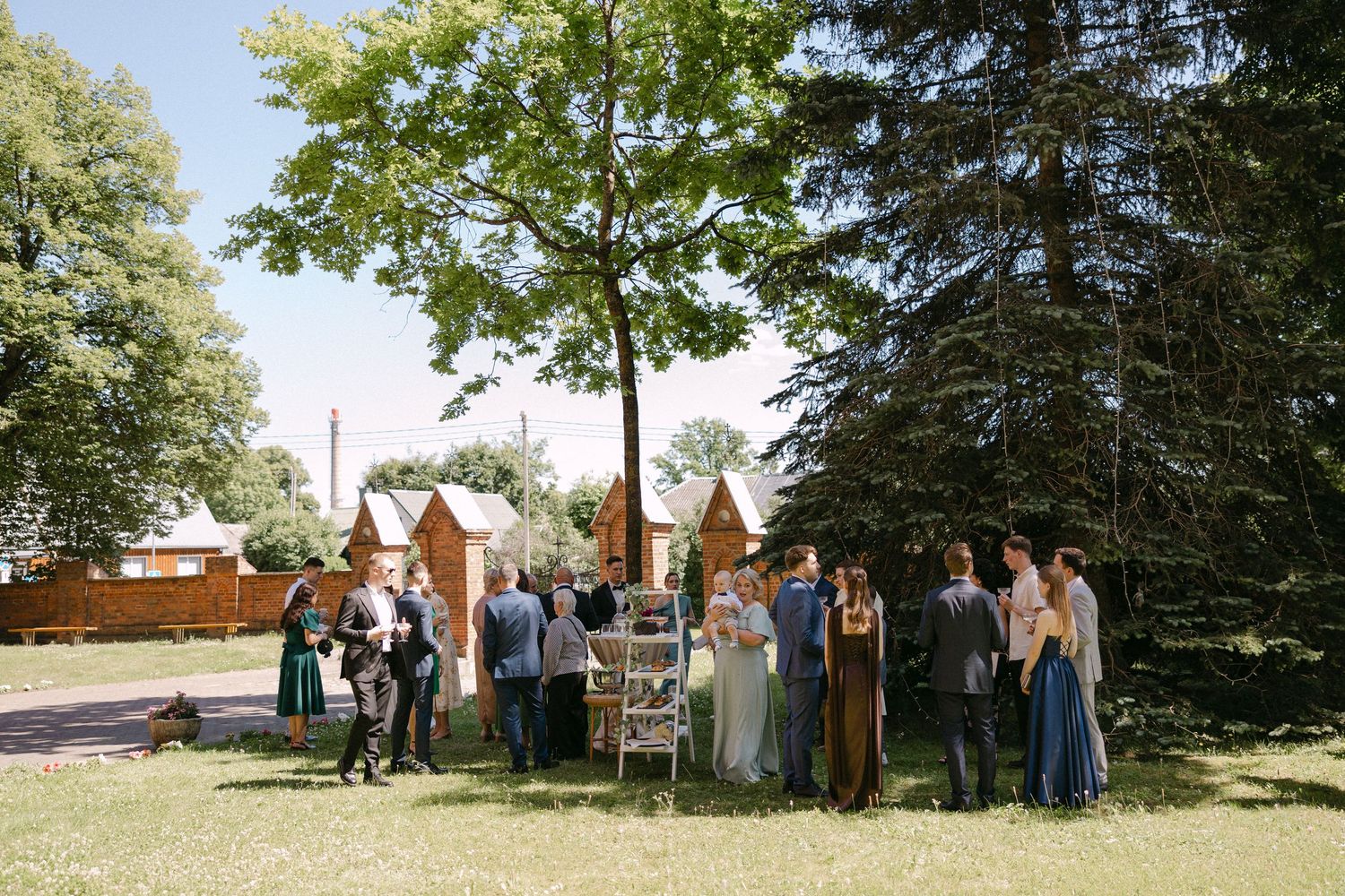 Wedding guests gather outdoors under large trees in a sunny garden during a summer wedding reception.