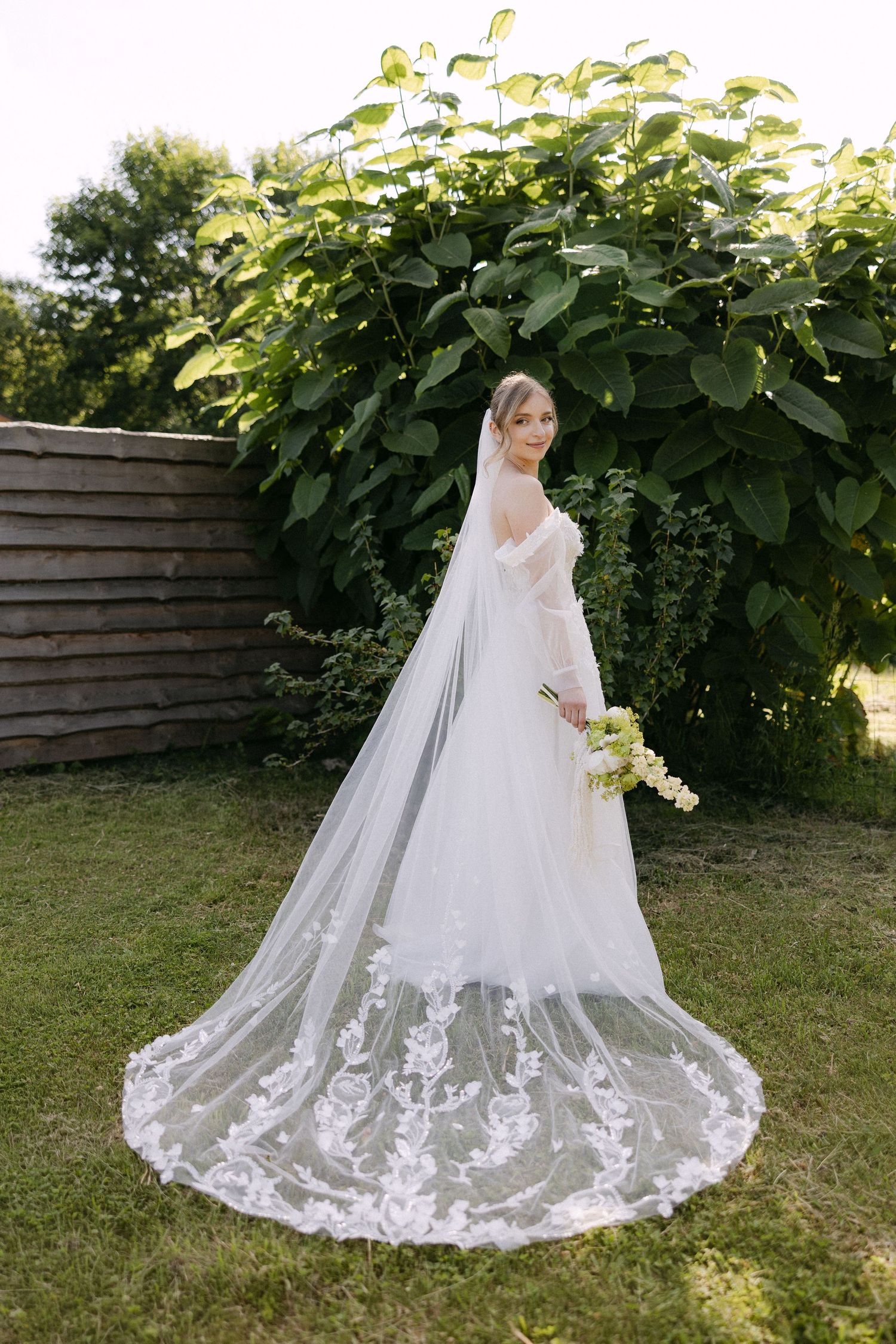 Bride in white off-shoulder gown with lace cathedral veil holds bouquet in garden setting.