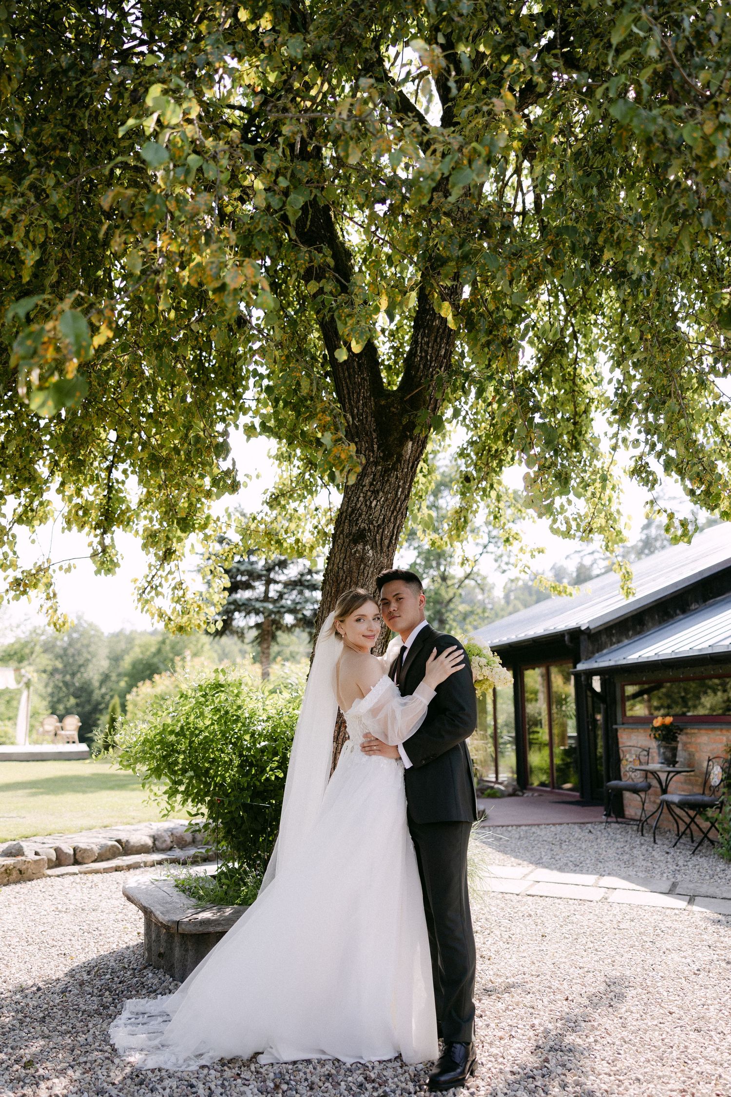 Bride and groom embracing romantically under a large green tree at an outdoor wedding venue.