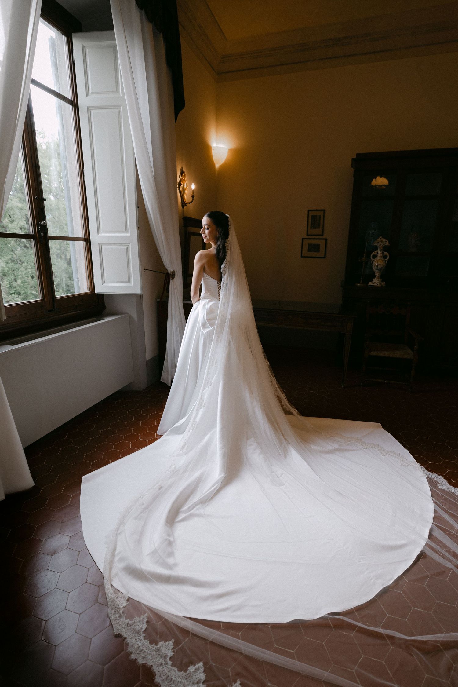 Bride in a stunning white gown with long cathedral veil standing by a window in an elegant villa.