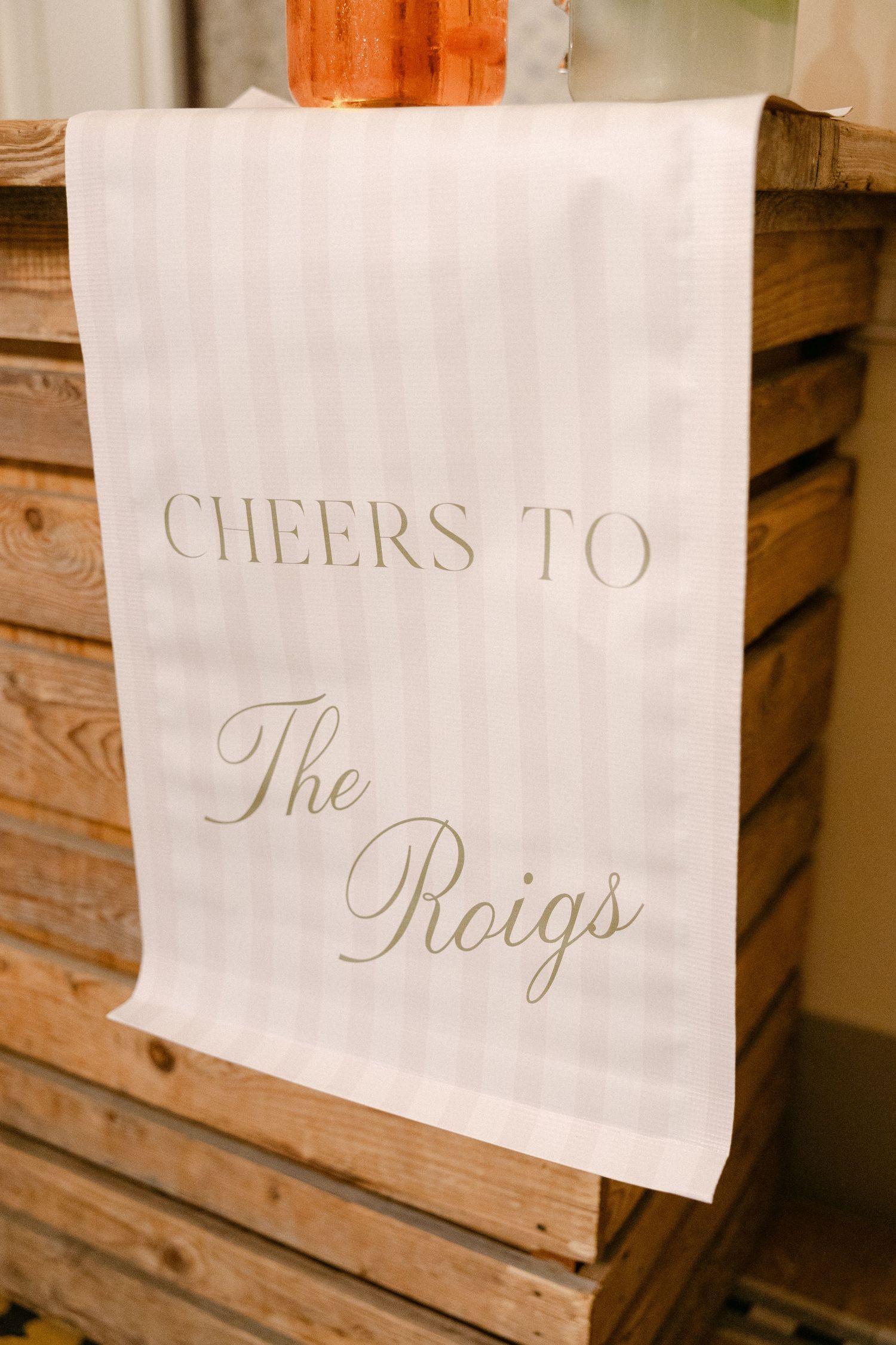 Elegant striped cloth bar sign reads Cheers to The Roigs in gold script on rustic wooden crate display.