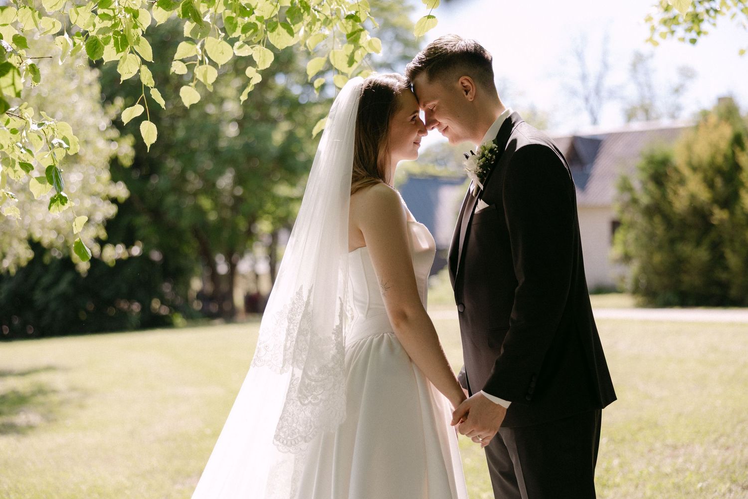 Bride and groom lean foreheads together holding hands in a sunny green garden on their wedding day.