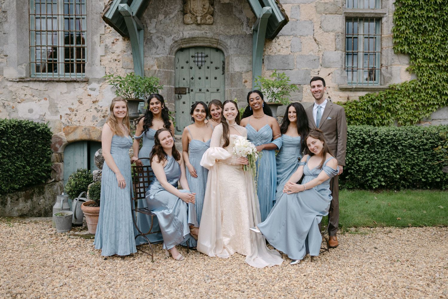 Bride in ivory gown poses with bridesmaids in dusty blue dresses before a rustic stone chateau.