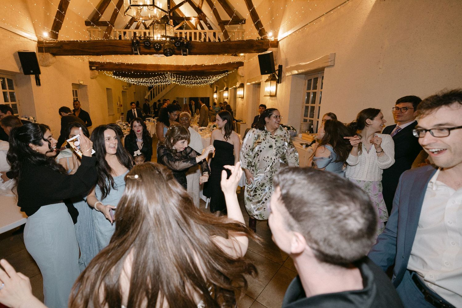 Lively wedding reception dance floor filled with guests celebrating under warm string lights in rustic barn venue.