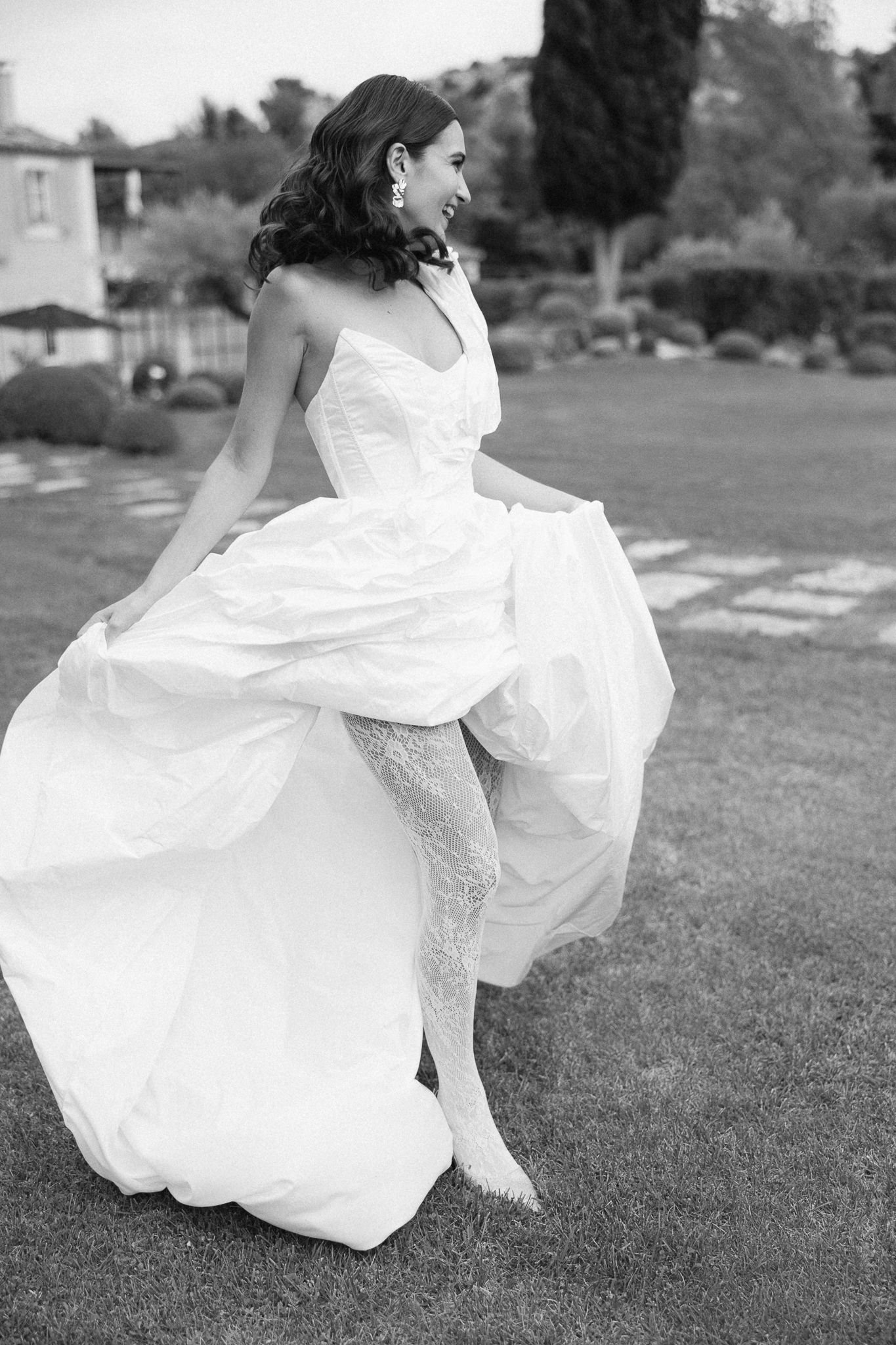 Black and white photo of a smiling bride in a flowing strapless wedding gown twirling on a lush garden lawn.