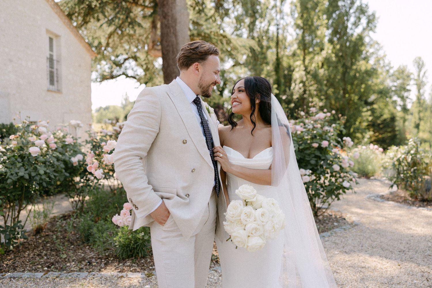 Bride and groom share a romantic moment outdoors, she holds white bouquet in strapless gown, he wears beige suit.