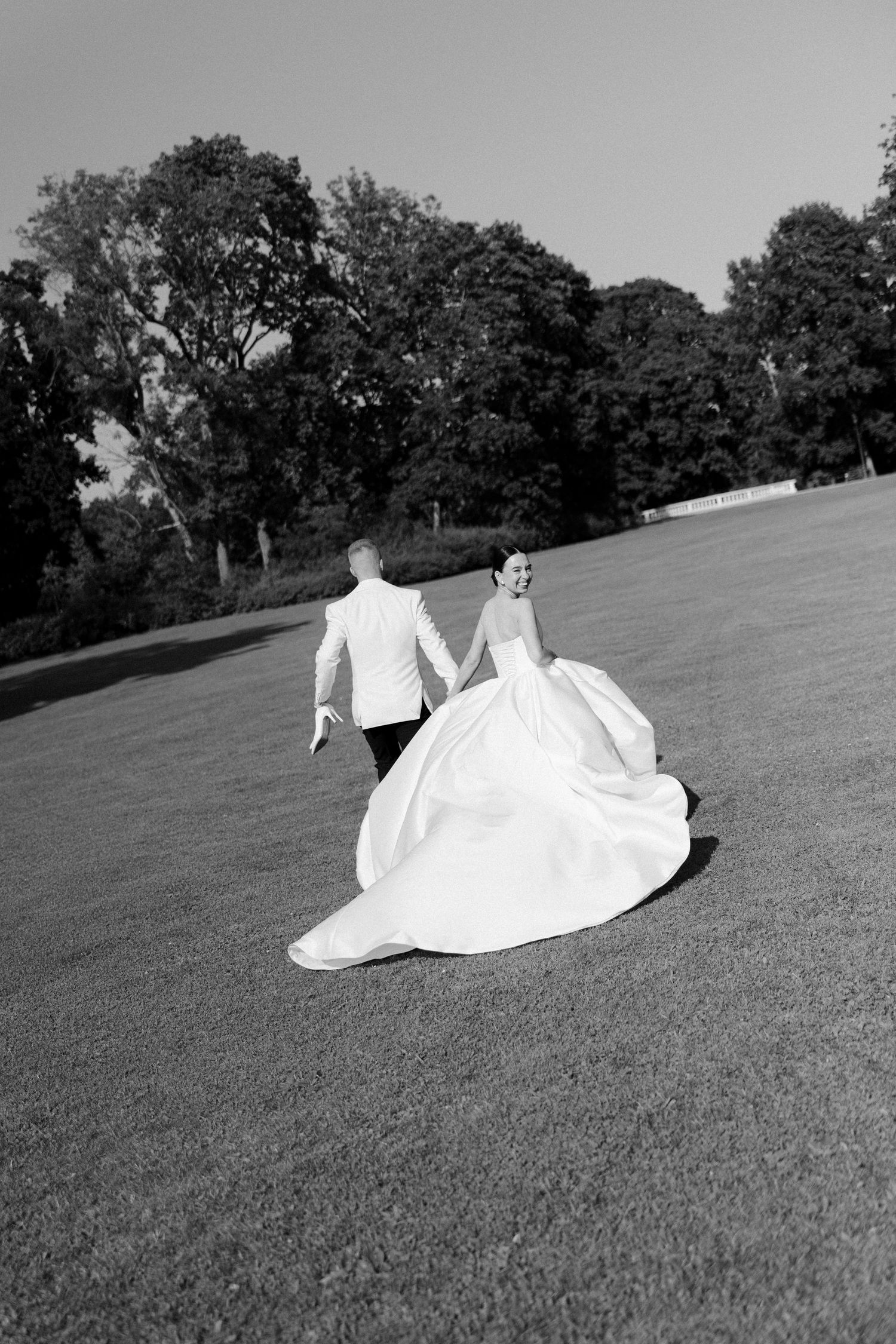 Black and white photo of bride and groom holding hands running across a lush green lawn with trees in background.