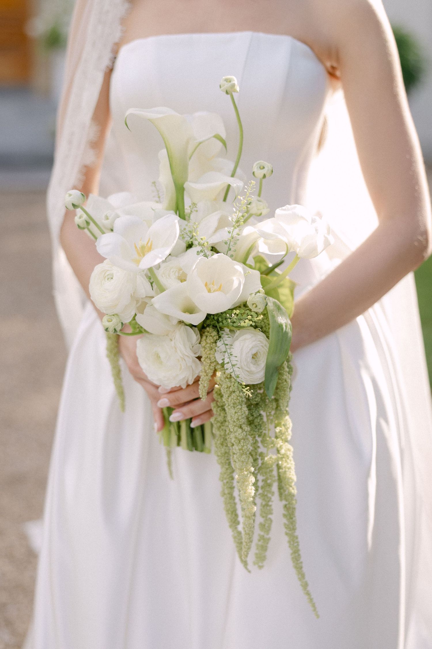 Bride holding elegant white wedding bouquet with calla lilies, ranunculus, and cascading green amaranthus.